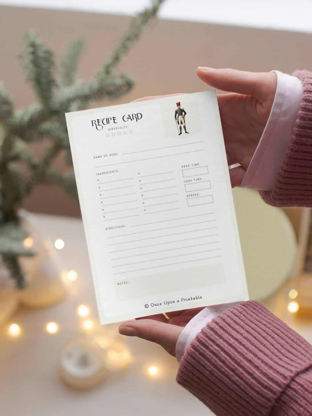 Nutcracker themed recipe card held by a woman wearing a pink sweater in front of a Christmas tree.
