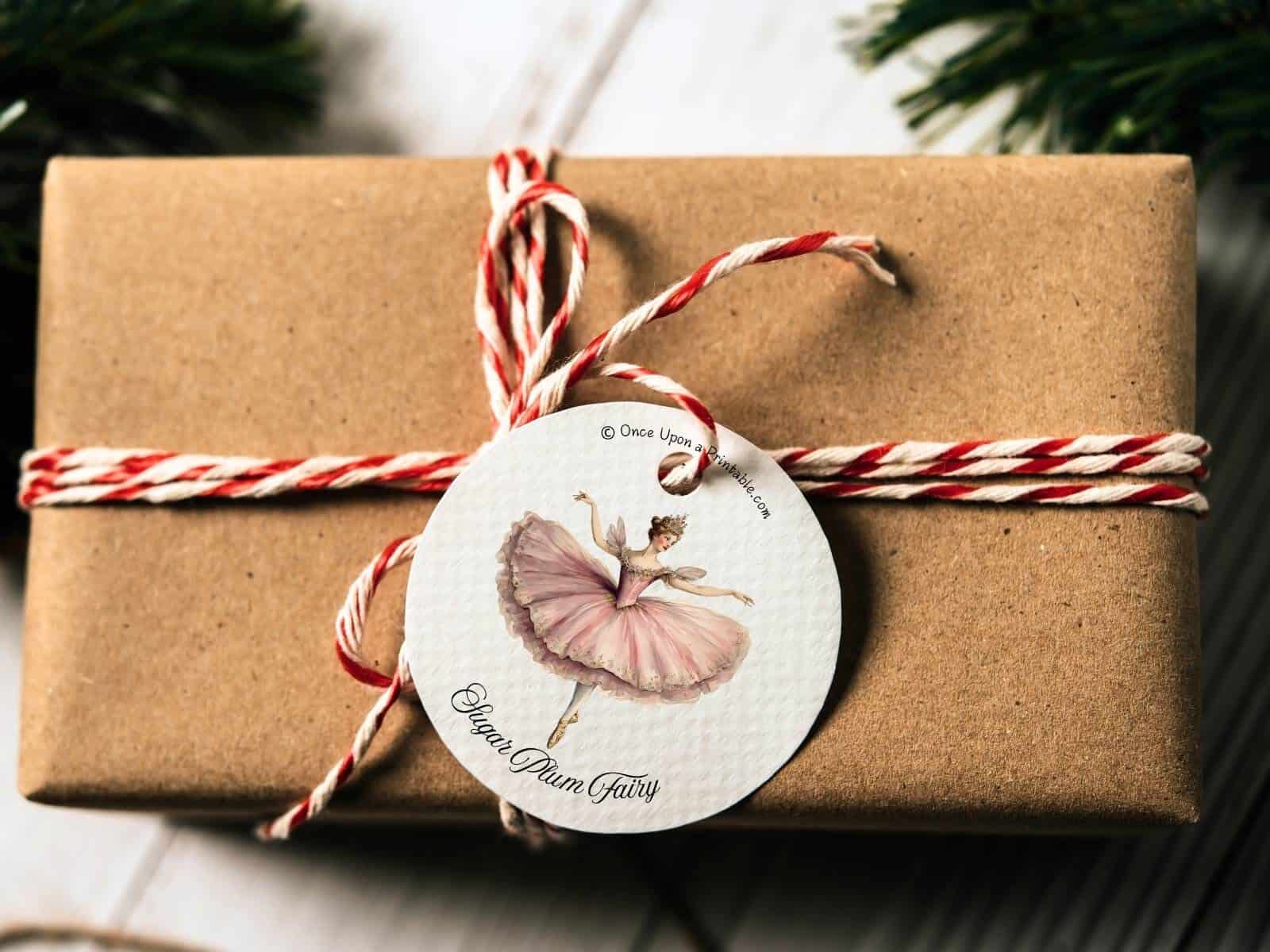 The Nutcracker themed gift tag with the "sugar plum fairy" ballerina atop a brown wrapped present with red striped string.