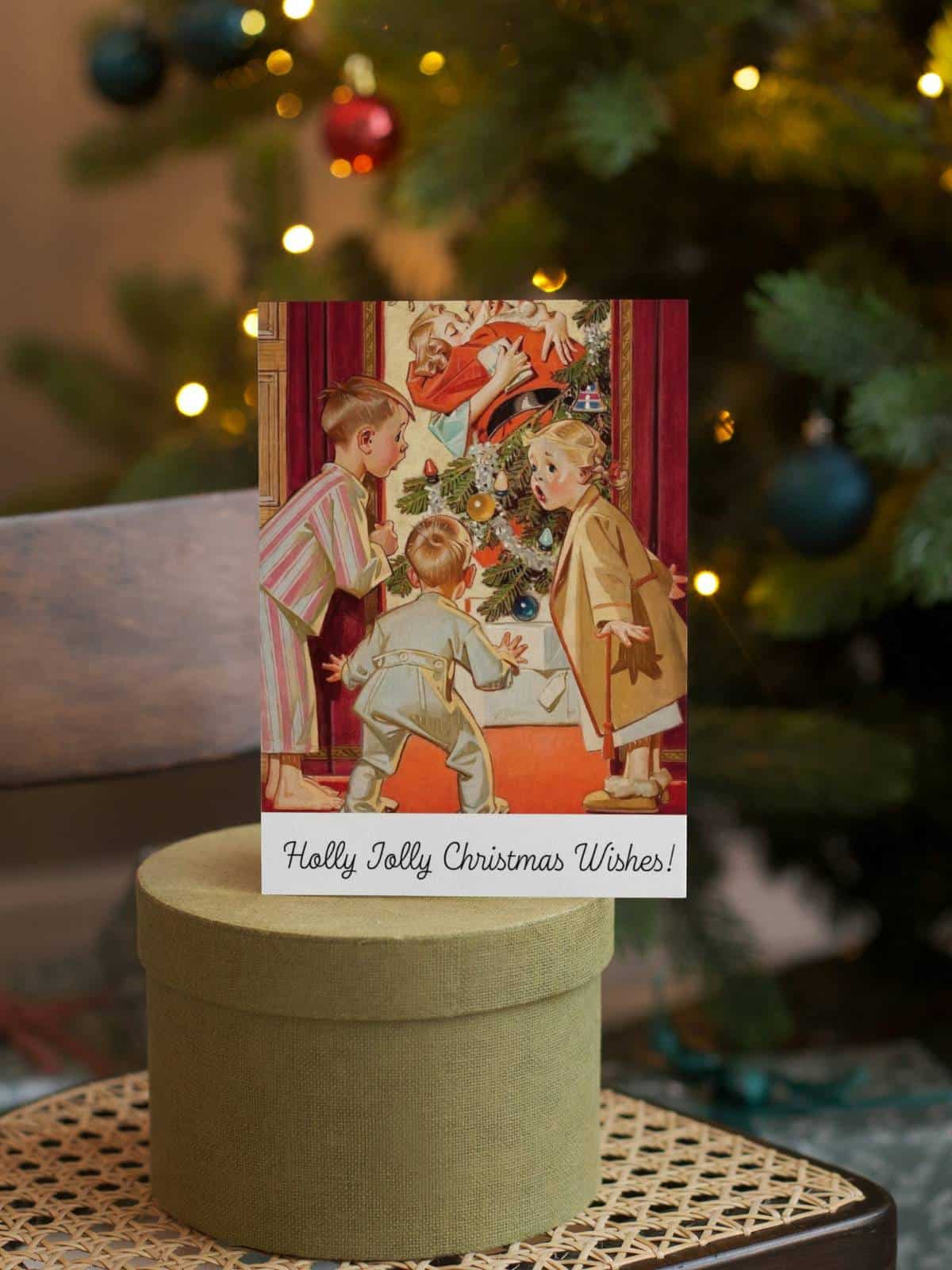 1920's vintage illustration of little children shocked watching Mommy kiss Santa Claus, a greeting card for CHristmas on display on top of a circular box in front of a Christmas tree. 