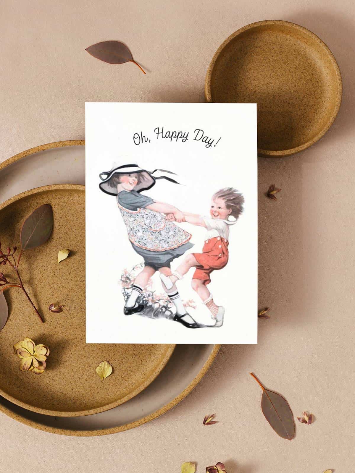 Children twirling together in the wind illustrated by Sarah Stilwell-Weber on a greeting card sitting on pottery plates with dried leaves.