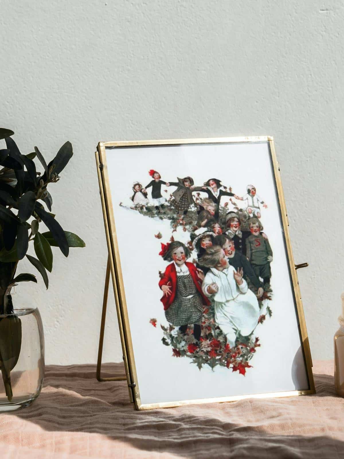 An illustration of children walking together in the fall leaves, framed and sitting on a surface next to a houseplant.