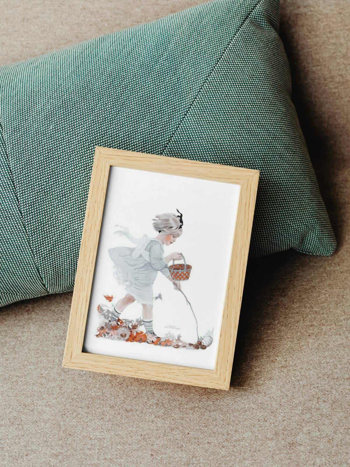 An illustration of a little girl walking in the fall leaves with a stick and a basket as the wind is blowing, framed sitting on a green pillow on the floor.