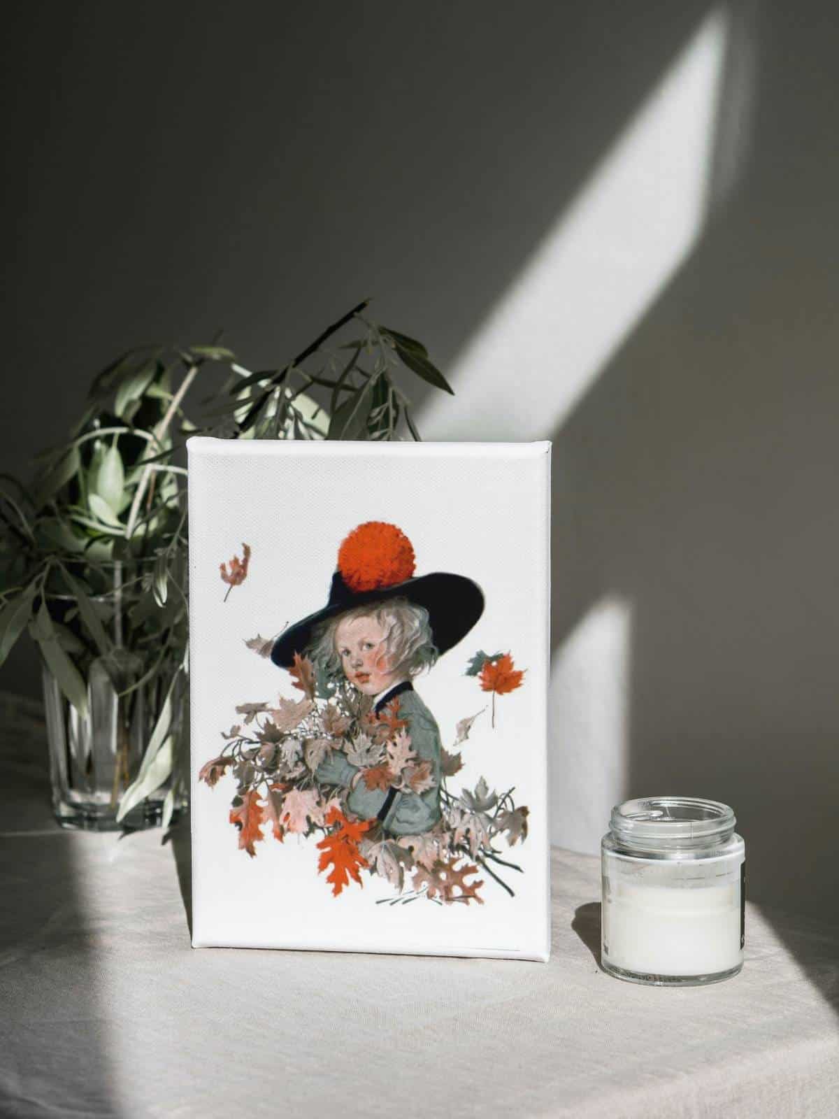 An illustration of a little girl wearing a hat with a large orange puff standing in fall foliage, framed sitting on a surface with a candle and a houseplant.