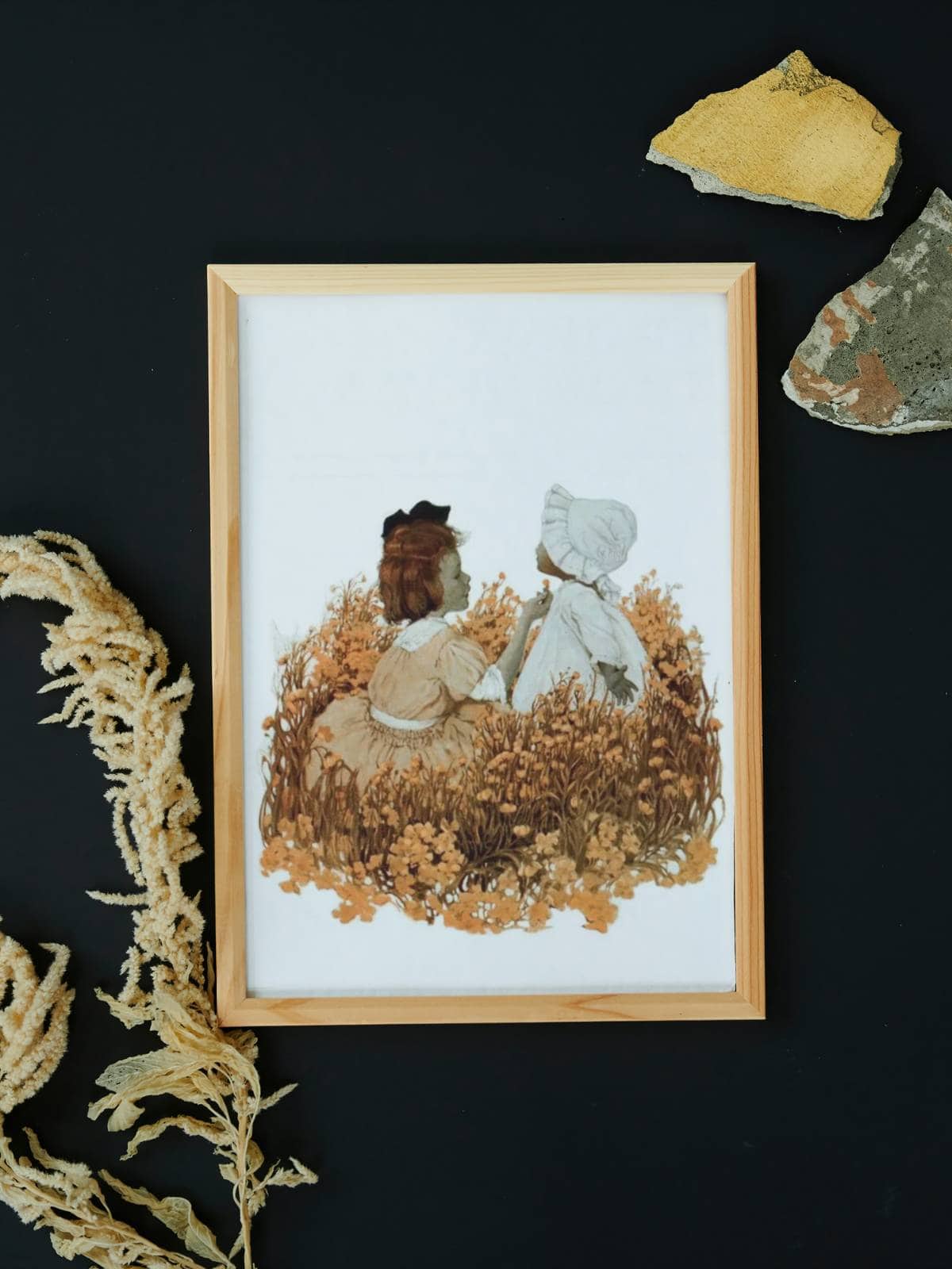 An illustration of two girls sitting in a fall meadow, framed sitting on a dark background with dried plants and rocks.