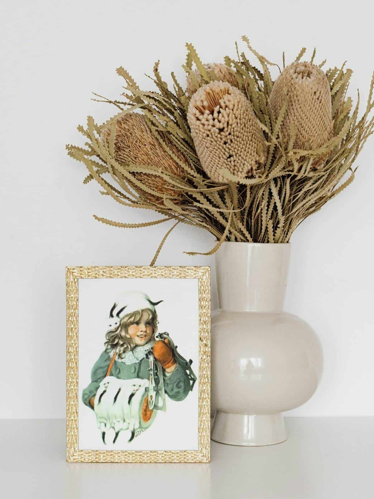 Winter illustration framed in a golden frame sitting next to a vase with dried flowers in it. 