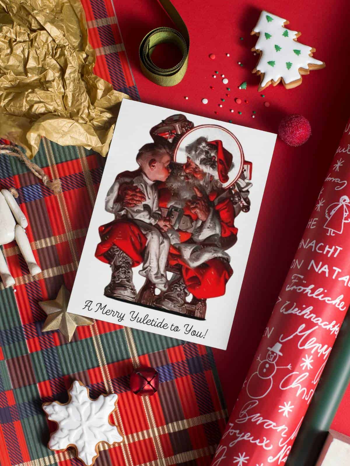 Santa Claus with a little boy sitting on his lap, a greeting card displayed on red Christmas wrapping.
