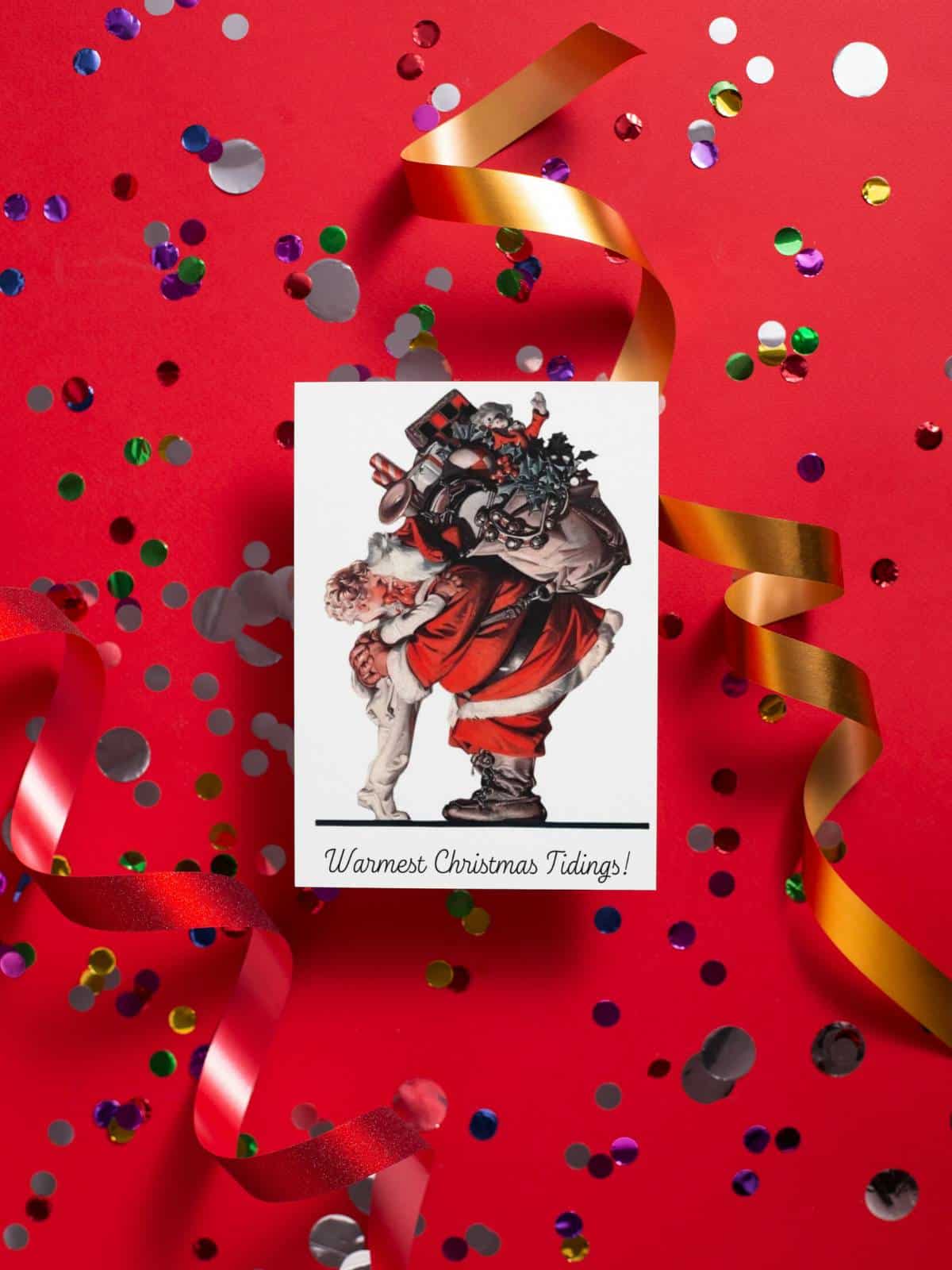Santa Claus giving a little boy in pajamas a hug, greeting card displayed on a red background with confetti and ribbon. 