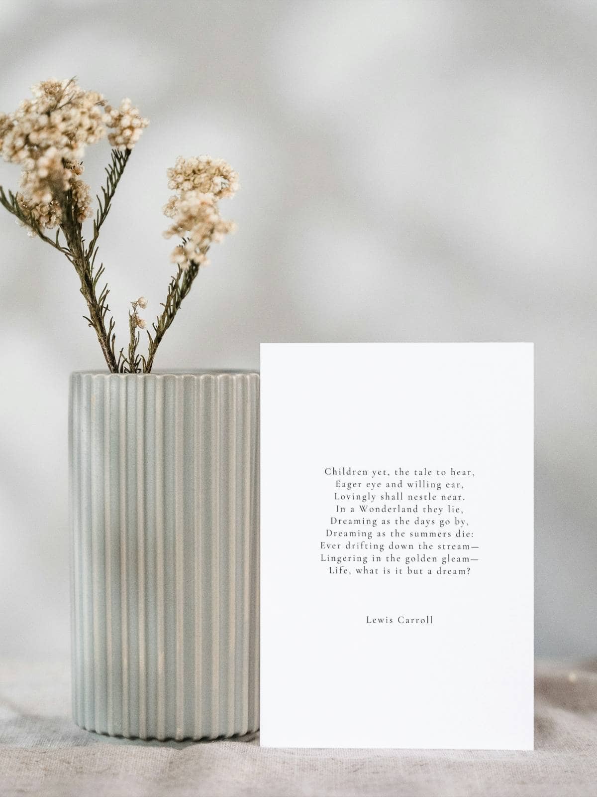 A poem by Lewis Carroll on a greeting card sitting next to a pottery vase with a dried flower sprig.