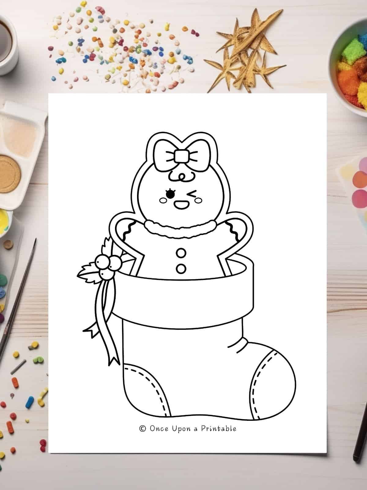 A winking baby gingerbread person sitting in a stocking, toddler coloring page with coloring supplies.