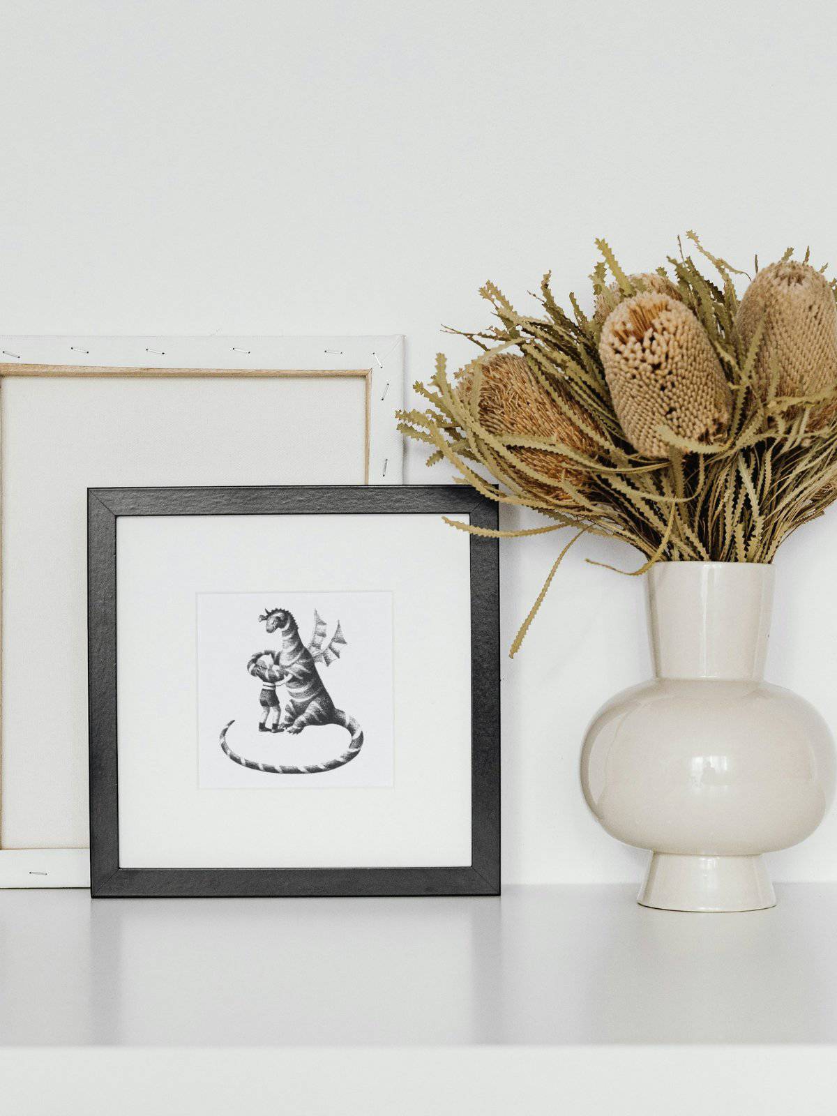 Black and white lithograph of Elmer and his Dragon friend, framed and displayed next to a vase of dried flowers. 
