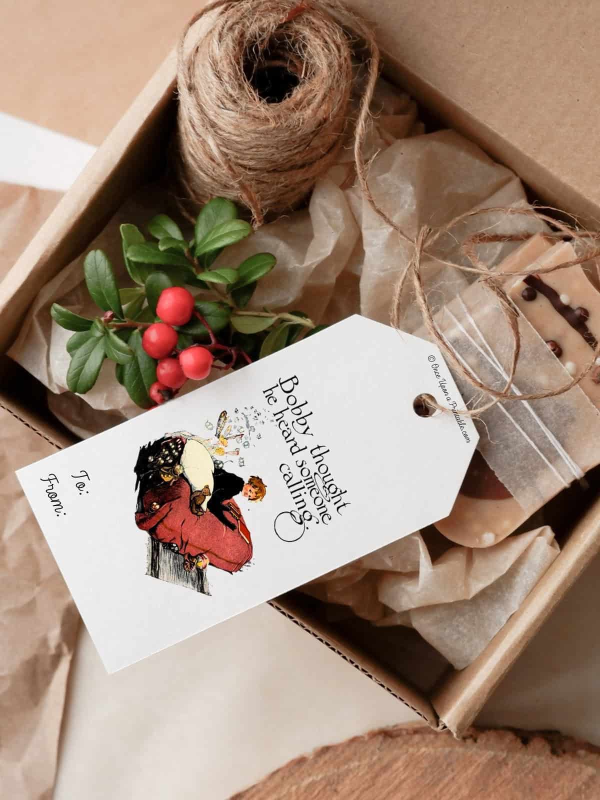 The Little Gingerbread man illustrated gift tag, full color, on top of a small box of gifts, a sprig of holly, and a coil of twine.