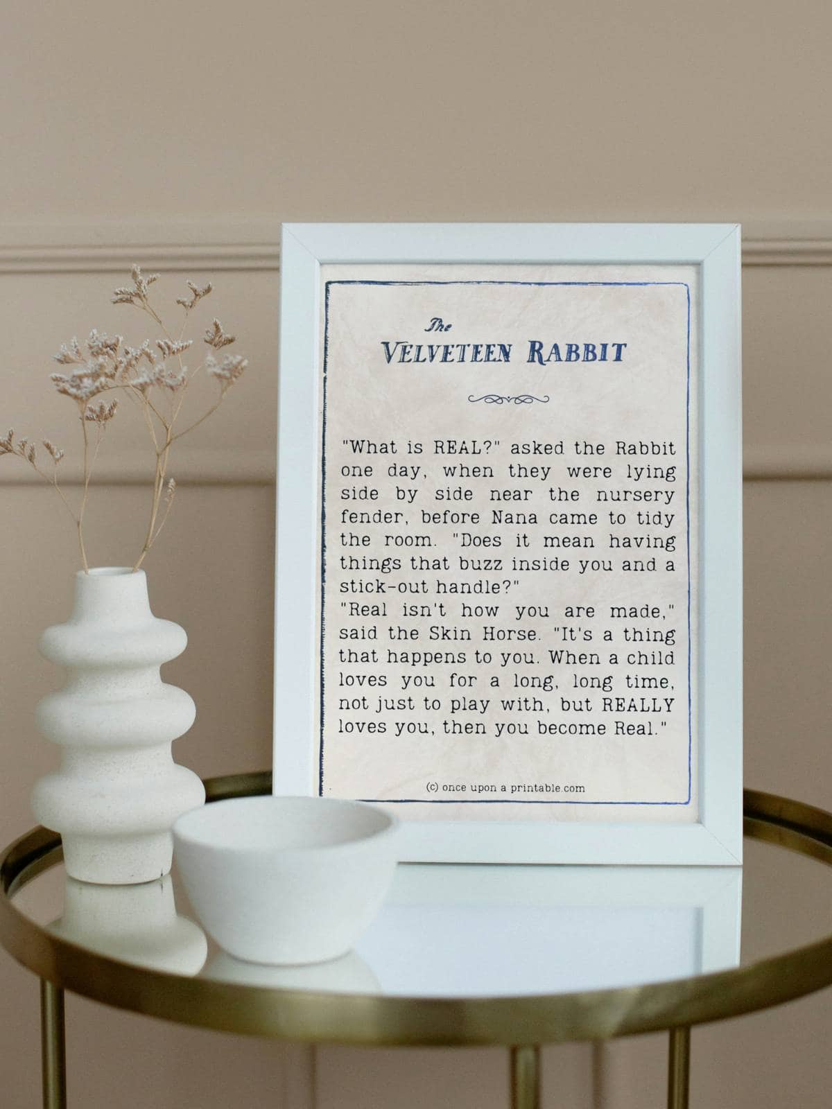 A quote from the book the velveteen rabbit framed in a white frame next to two white vases on a glass table.