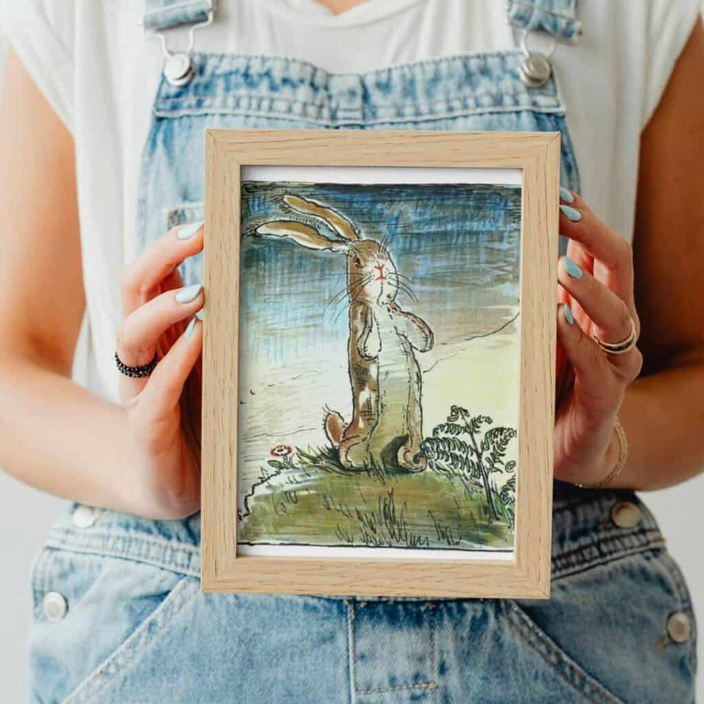 A vintage illustration of the Velveteen Rabbit in full color framed and being held in a woman's hands that is wearring coveralls and a white t shirt.