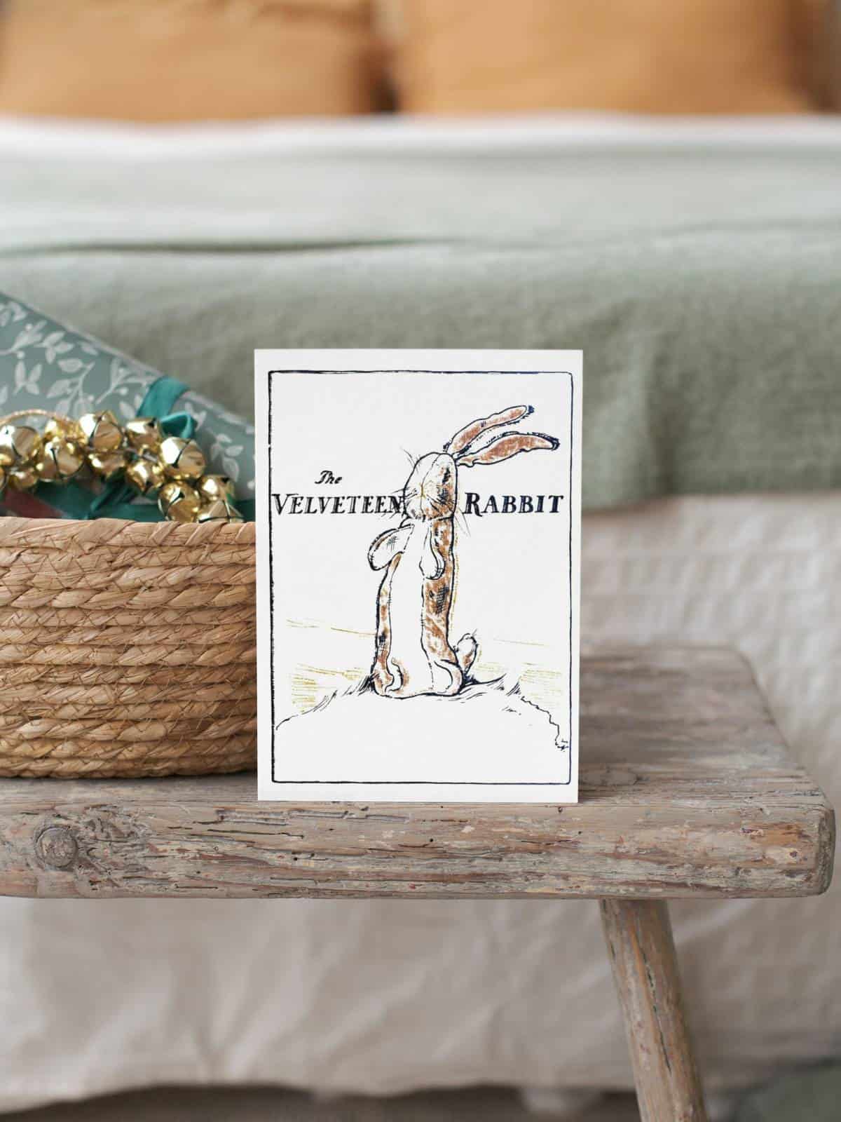 The Velveteen rabbit original illustration on a note card sitting on a wooden bench next to a bed and basket.