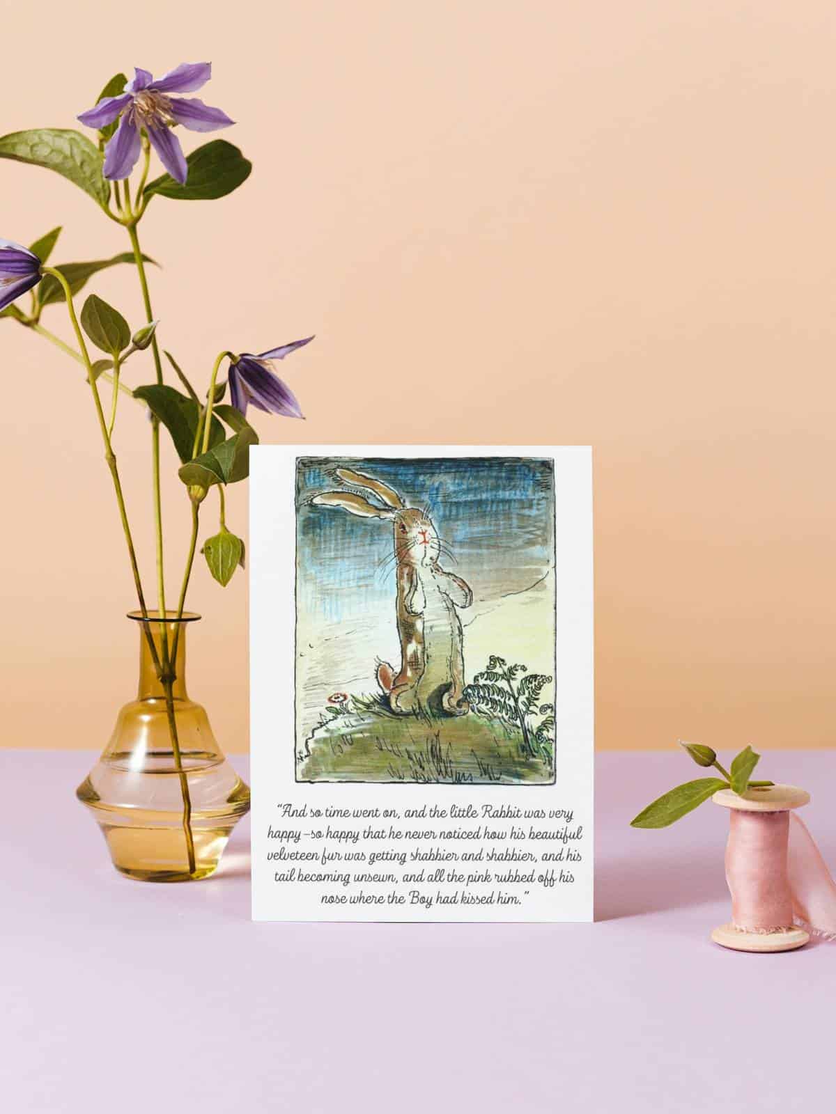 An illustrated Velveteen Rabbit note card in full color displayed with a vase and flowers and a spindle of ribbon.