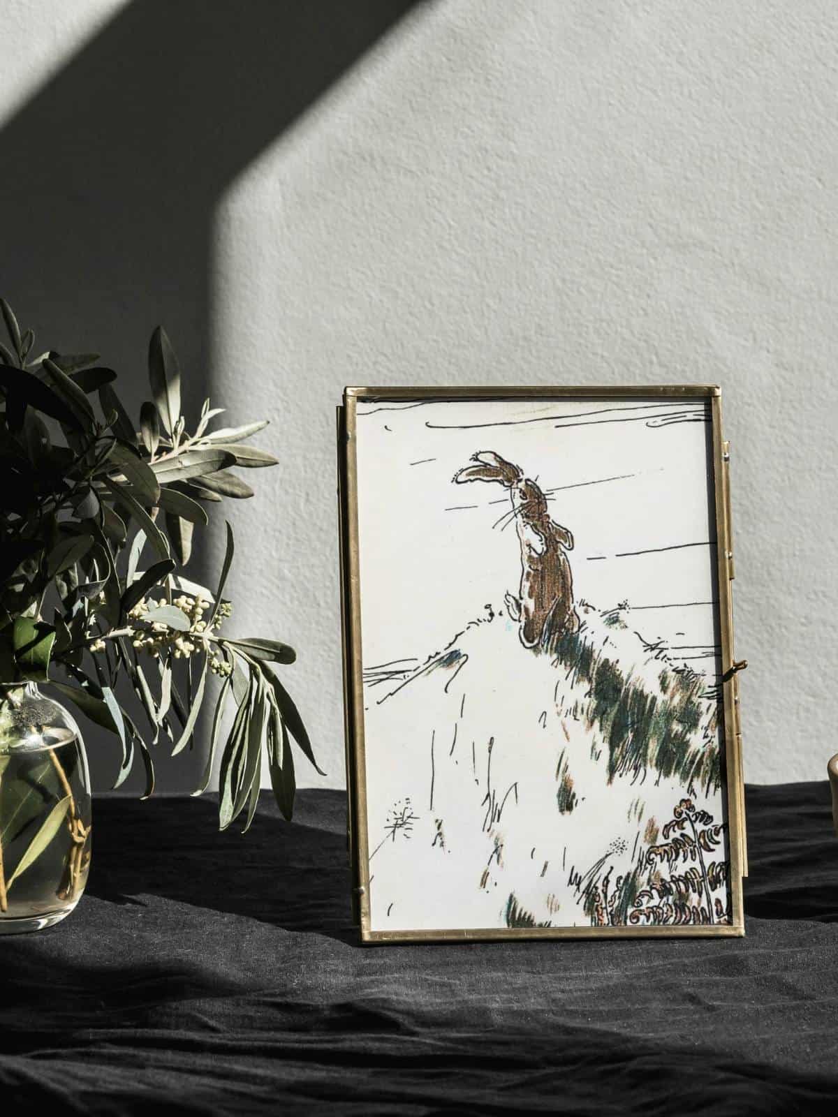 The Velveteen Rabbit sitting on a hill watching real rabbits play, illustration from the book framed and displayed next to a house plant.