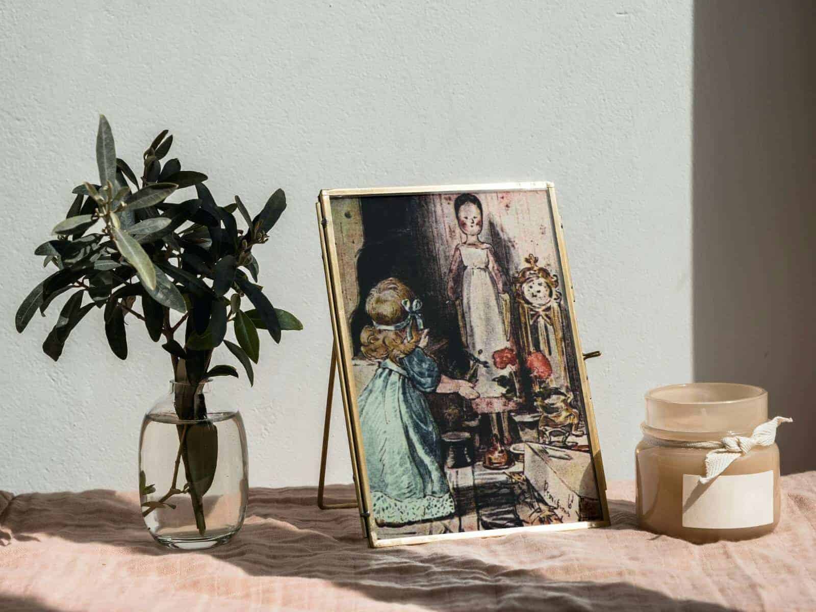 Dolls in their dollhouse illustrations by Beatrix Potter framed and displayed next to a candle and a bud vase with a few sprigs.