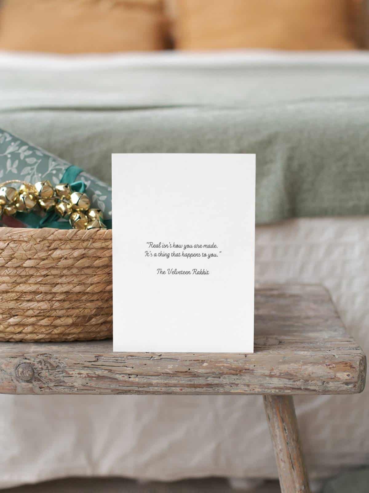 The Velveteen rabbit original illustration on a note card sitting on a wooden bench next to a bed and basket, the inside with a quote.