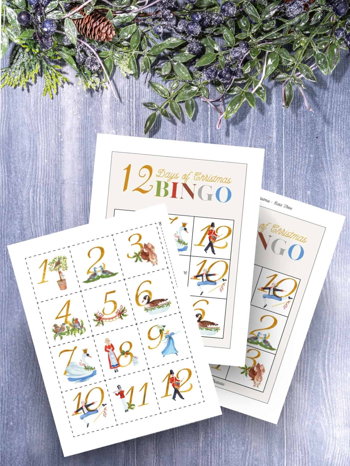 12 Days of Christmas bingo cards and calling cards printed and laying on a wooden surface with Christmas holly.
