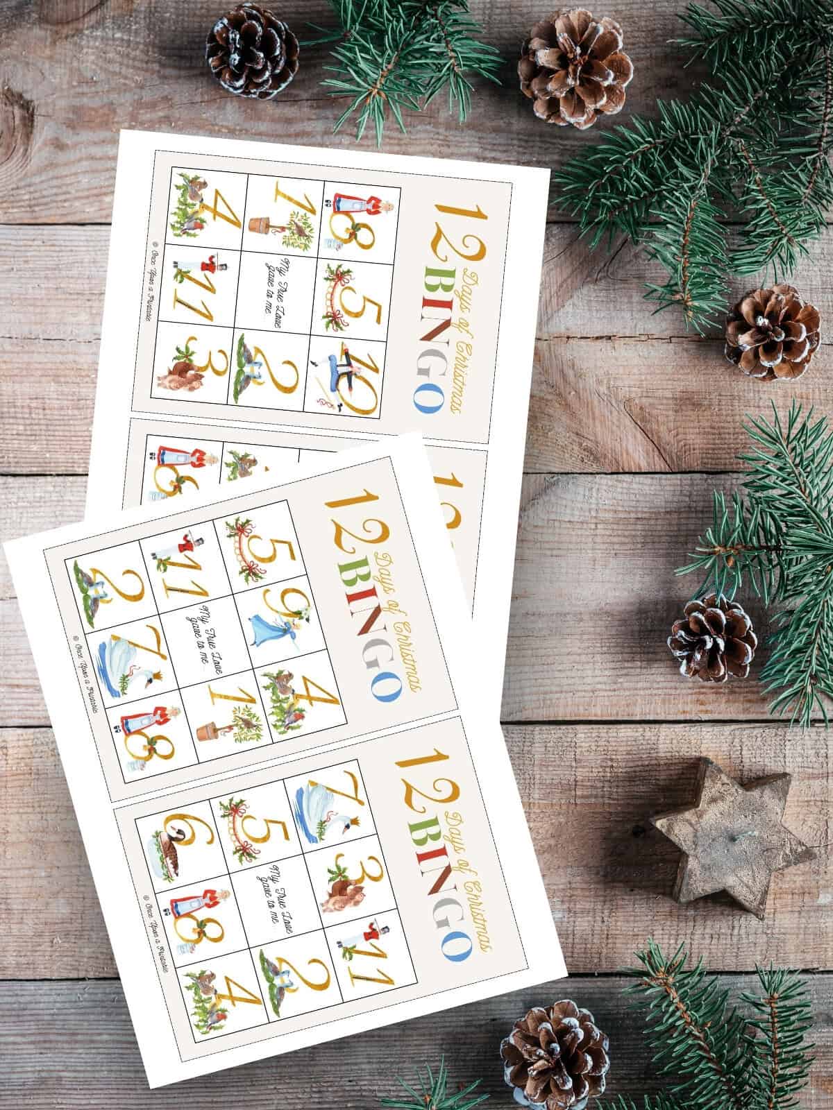 12 Days of Christmas bingo cards with watercolor graphics printed and laying on a wooden surface with garland.