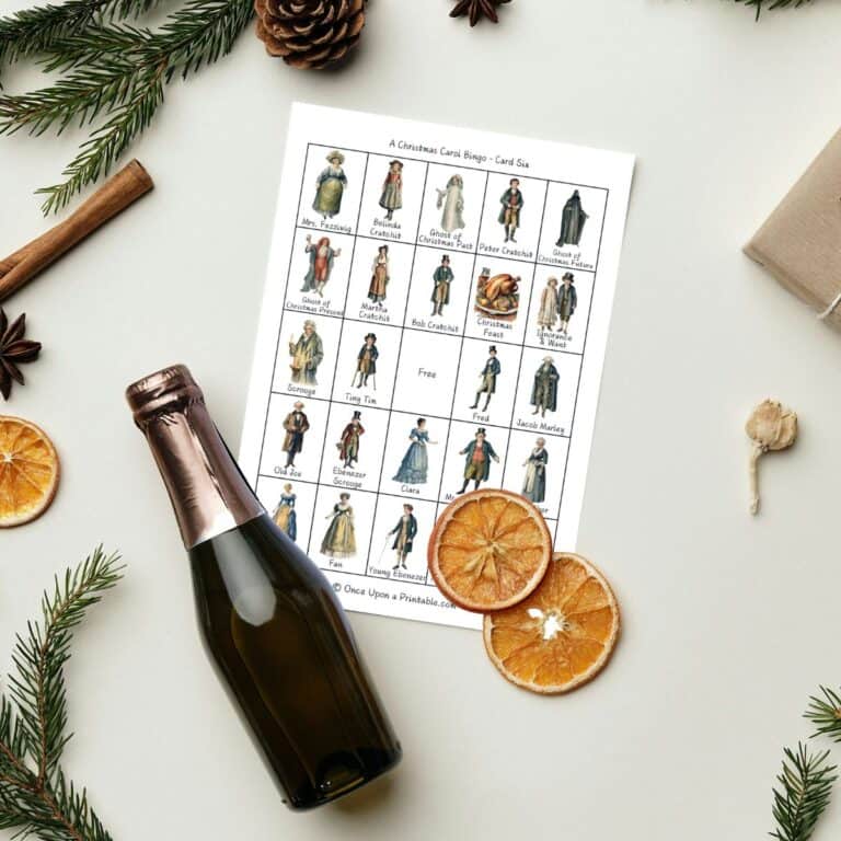 A charles dickens inspired "a christmas carol" bingo game printables laying on a white table with dried orange slices, a bottle of apple cider, garland, and cinnamon sticks.