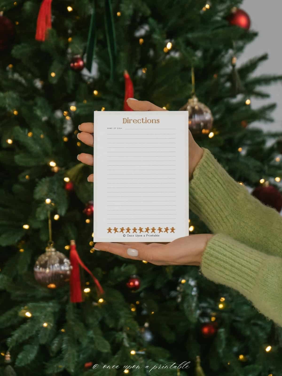 The back of a watercolor graphic designed recipe card with gingerbread men on it being held in the hands of a woman in front of a Christmas tree. 