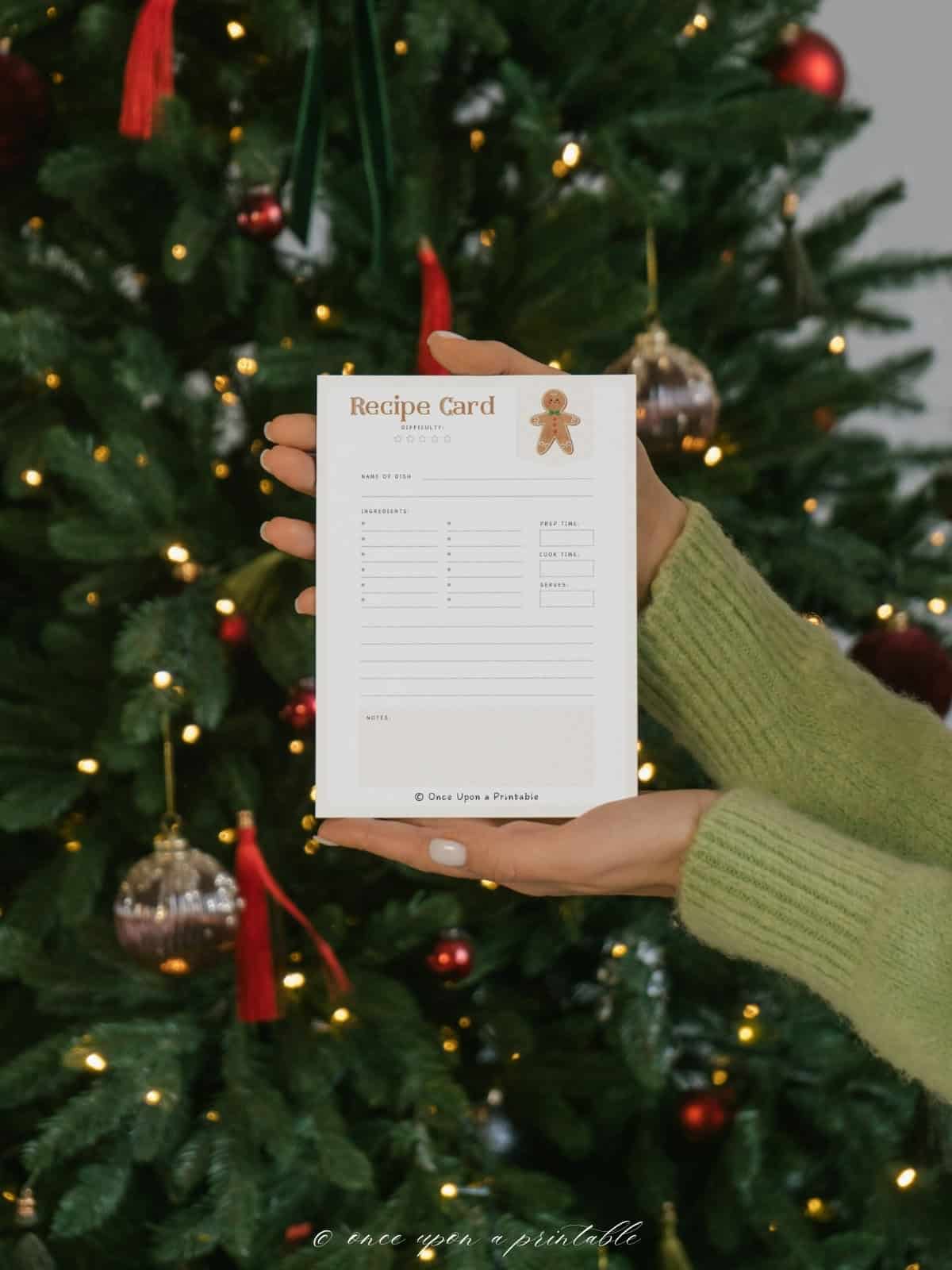 The front of a watercolor graphic designed recipe card with gingerbread men on it being held in the hands of a woman in front of a Christmas tree. 