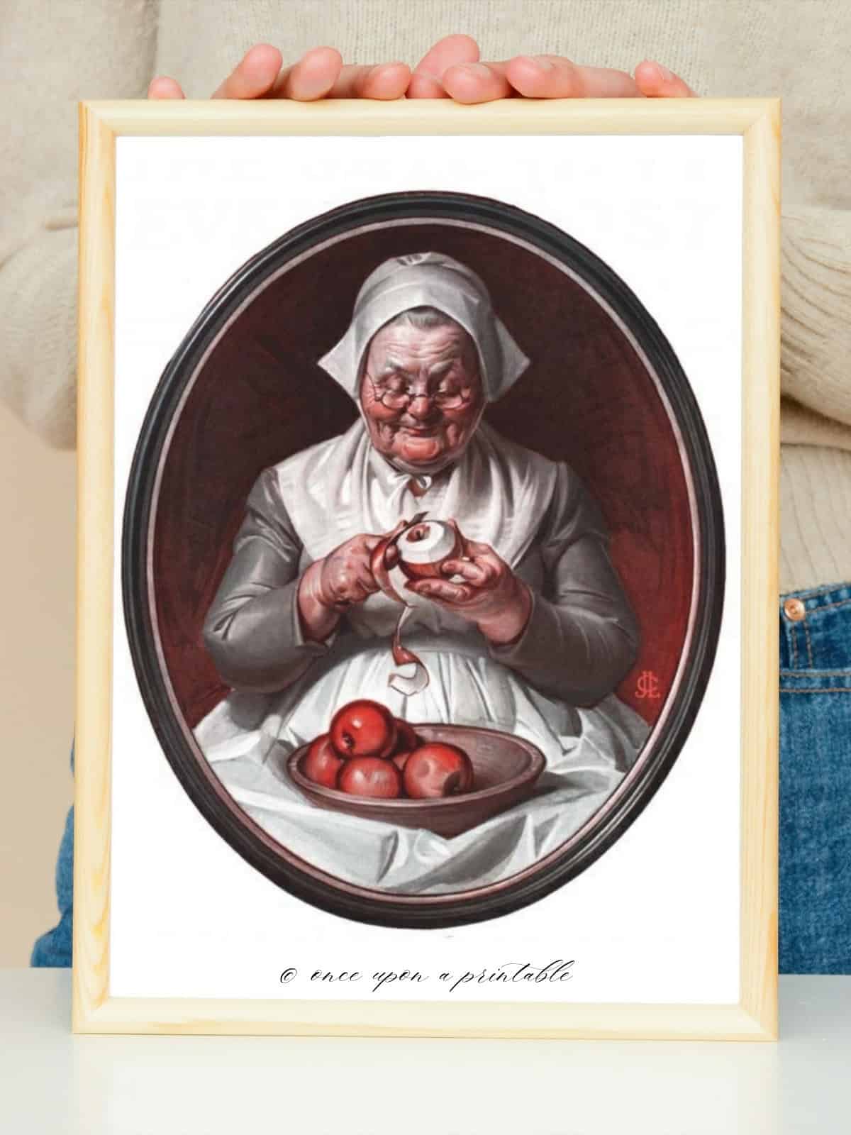 A vintage illustration of a grandmother pilgrim dressed woman peeling apples with a bowl of apples in her lap, framed and held by a woman's hands.