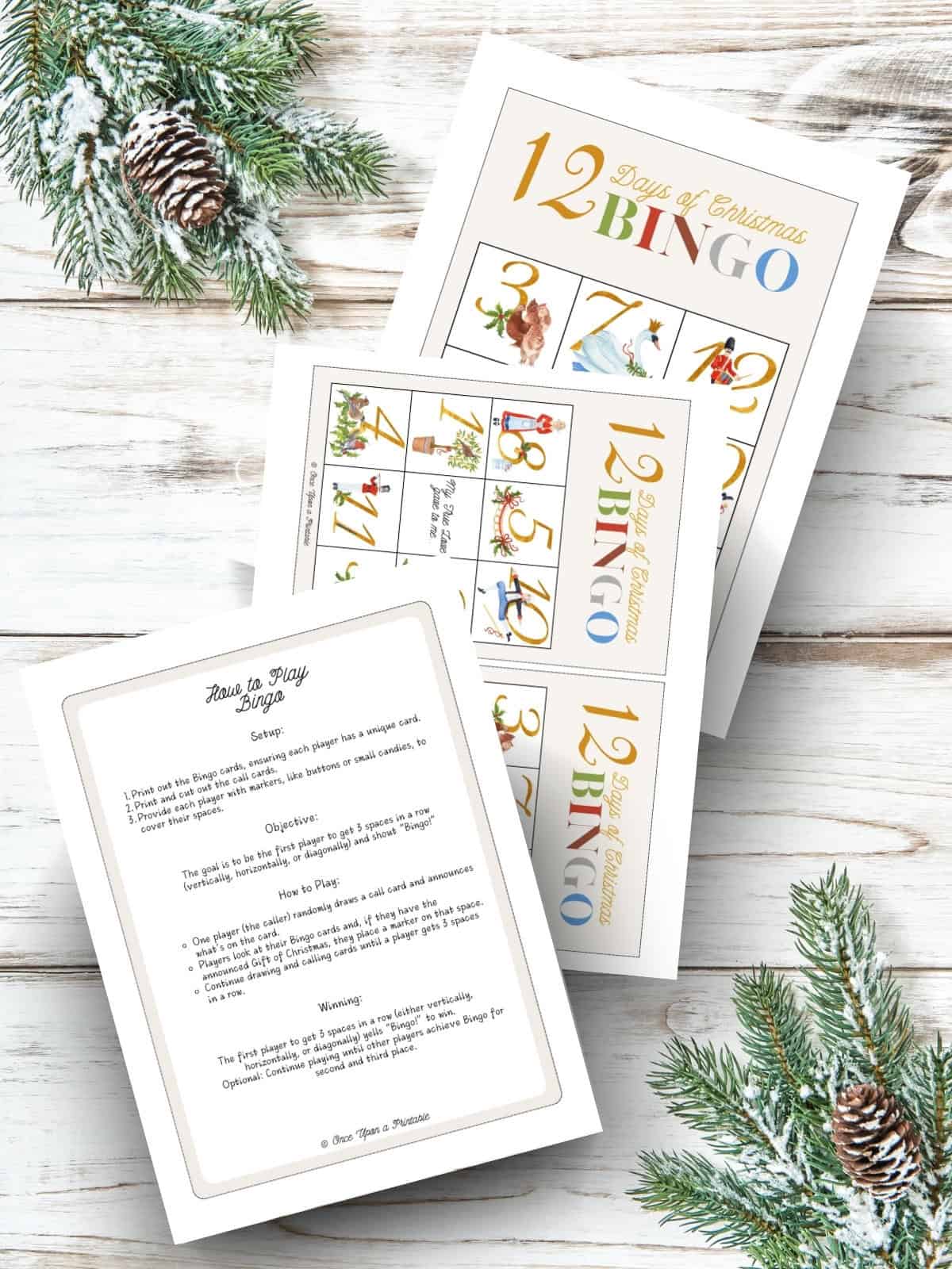 12 days of christmas themed bingo card game with instructions and bright watercolor illustrations printed and laying on a white-washed wooden surface with garland.