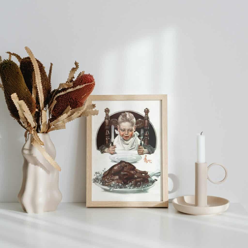 Vintage illustration of a boy excited to eat his turkey dinner, framed and displayed next to a vase with dried flowers and a candle stick.