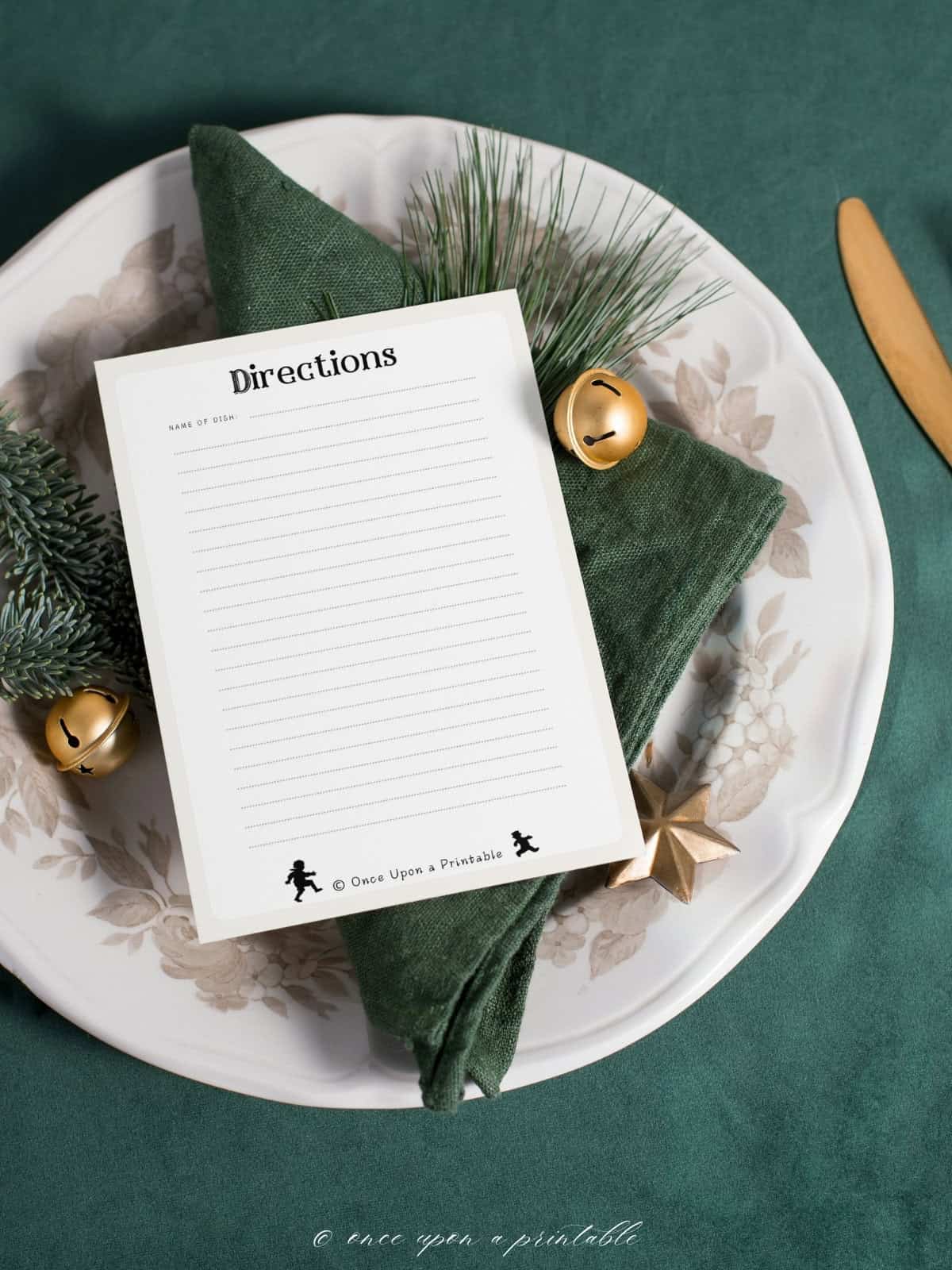 The back of a vintage illustrated black and white recipe card sitting on a beautiful Christmas place setting with green napkin and brass bells. 
