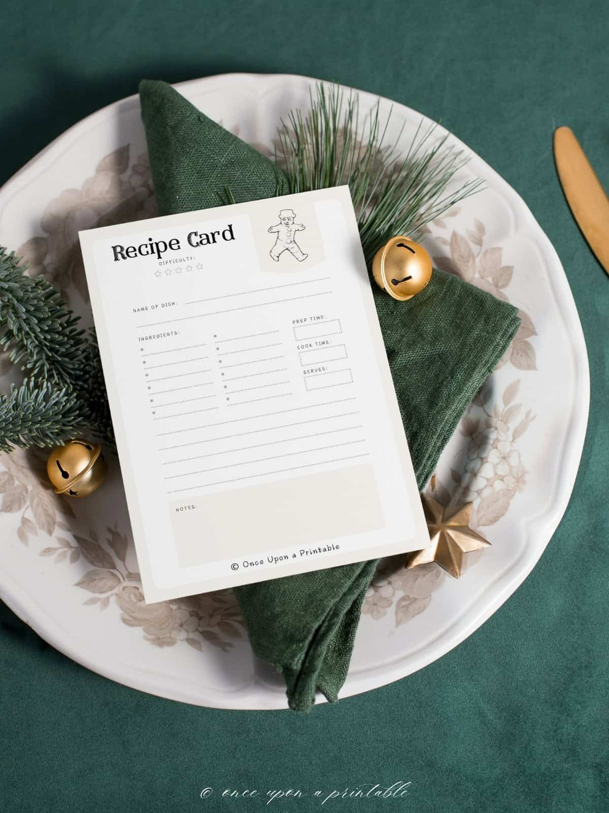 The front of a vintage illustrated black and white recipe card sitting on a beautiful Christmas place setting with green napkin and brass bells. 