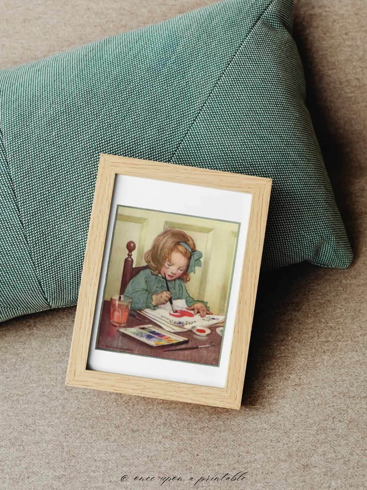Vintage illustration of a little girl painting, framed and laying on a green pillow.