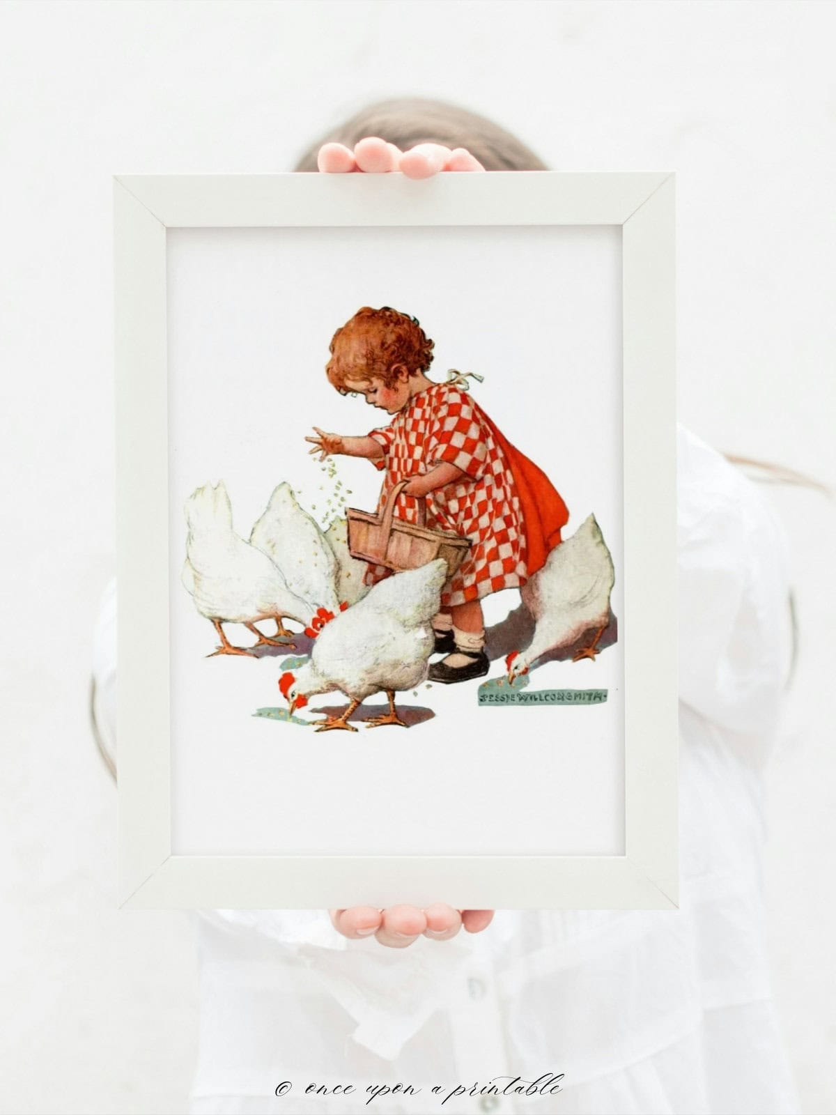 Vintage illustration of a child feeding chickens, framed in a white frame and held in a woman's hands.
