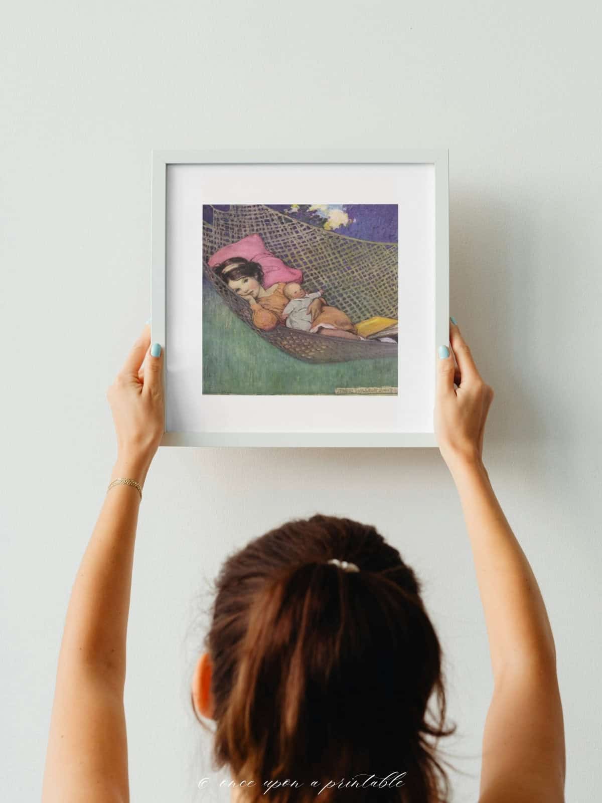 A Little girl swinging in a hammock with her baby doll, illustrated and framed held up by a woman's hand.