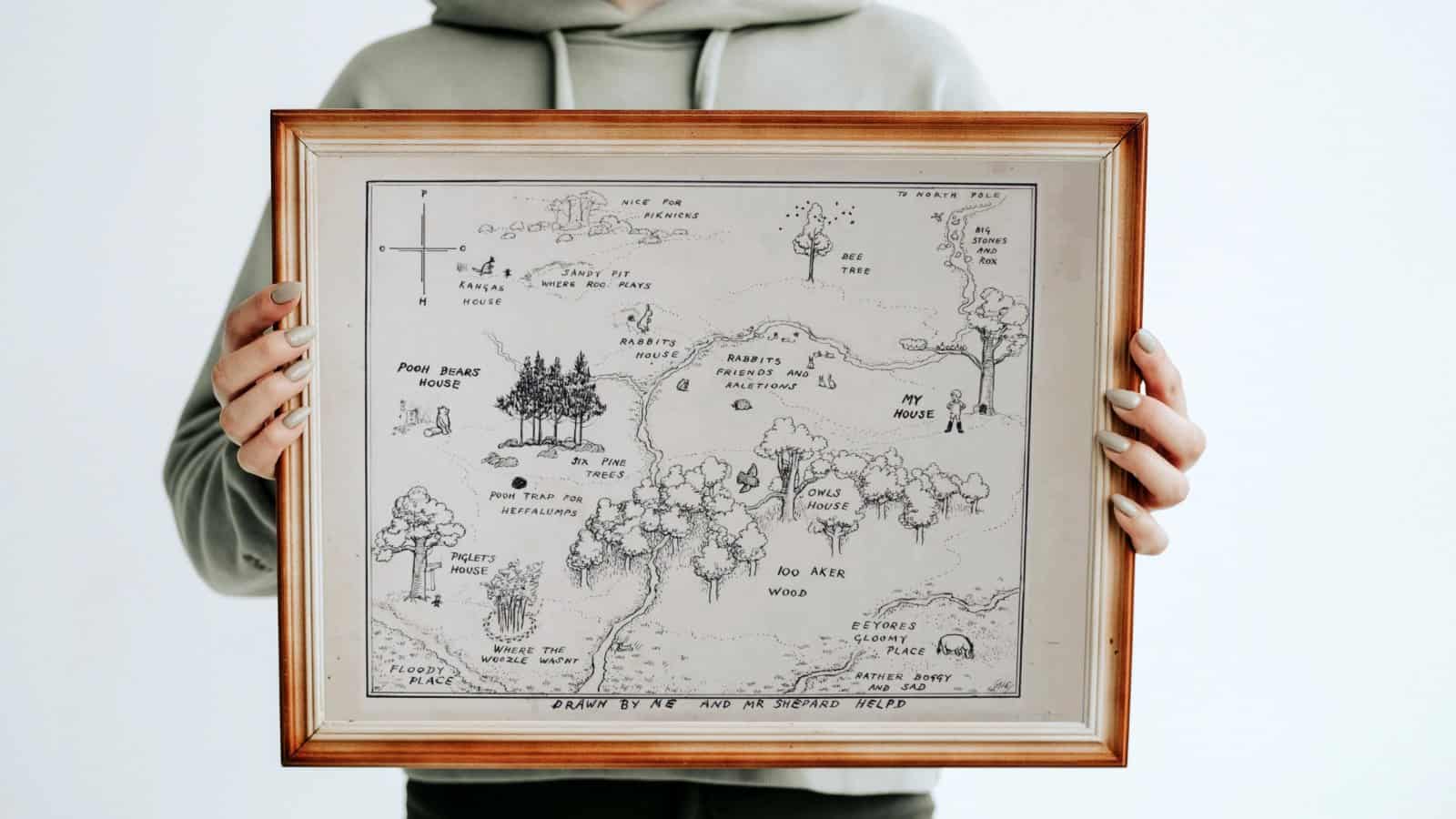 100 Acre wood map by E. H. Shepard and A. A. Milne, winnie the pooh and other beloved characters drawn on a map framed and held in a woman's arms.