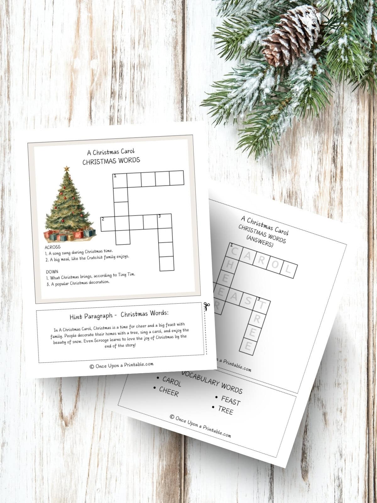 A Christmas Carol Crossword Puzzle and the answer keys laying on a wooden background with a garland sprig and pinecone.