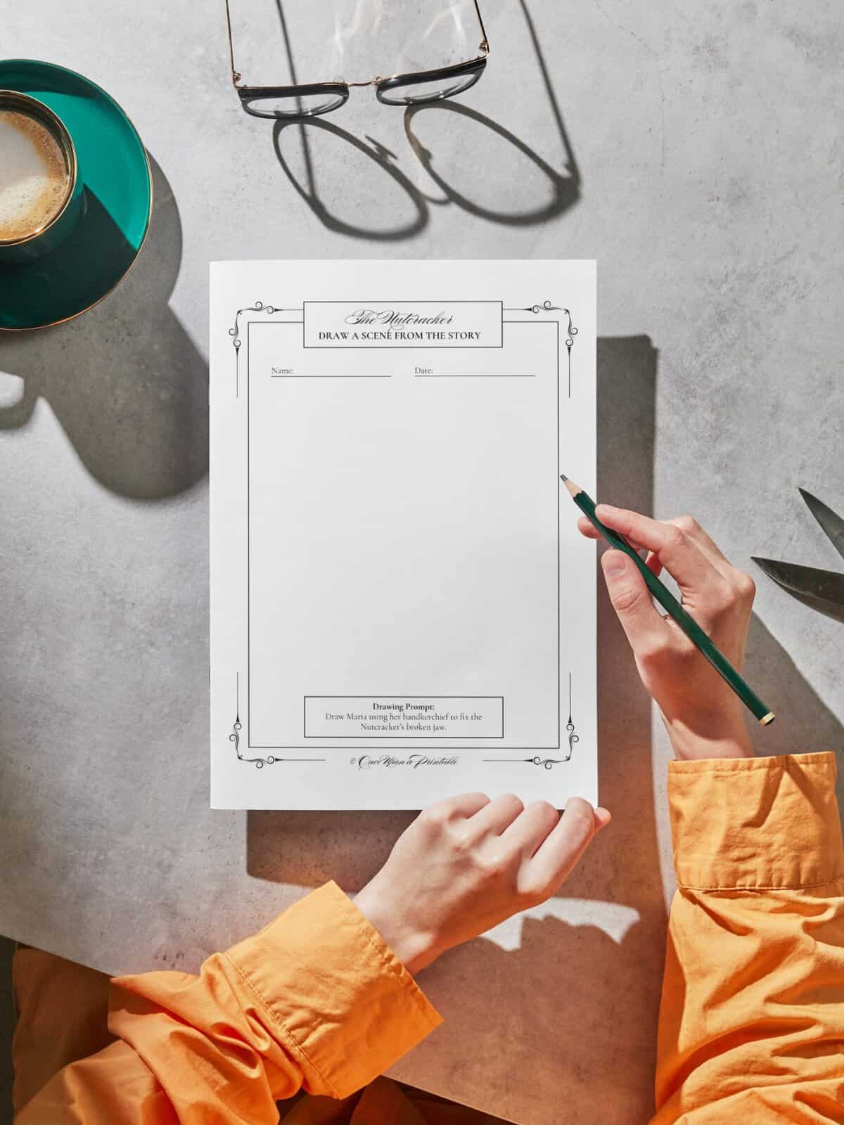 The Nutcracker and the Mouse King drawing prompt page being held in a woman's hand and her other hand holds a pencil, displayed with glasses and a cup.