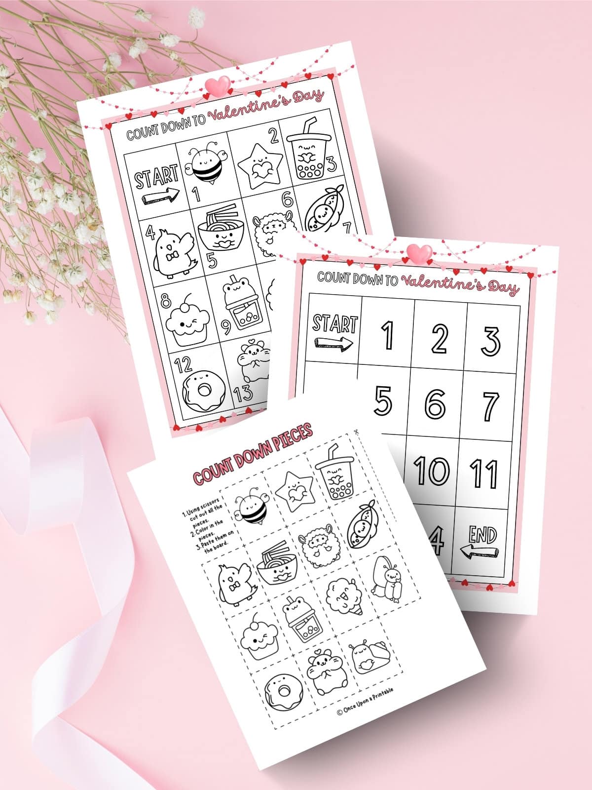 Two different interactive coloring countdown calendar's for Valentine's day with cute kawaii-style grahpics, laying on a pink background with baby's breath flowers and white ribbon.