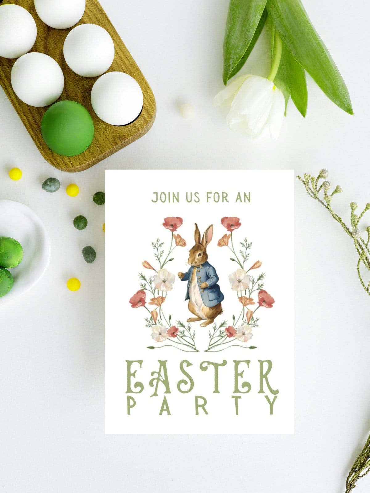 Easter Party invitation card with peter rabbit on the front laying on a white table with eggs and tulips.