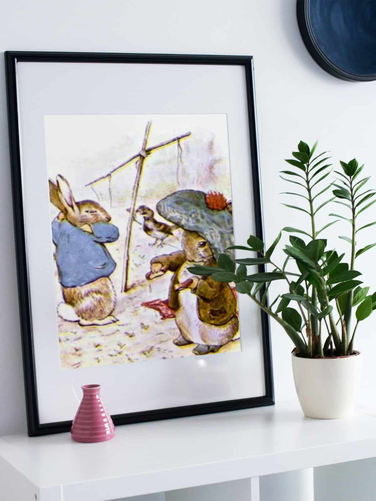 Benjamin Bunny illustration with Peter Rabbit in Mr. McGregor's garden, framed and displayed on a white console table next to a potted plant.