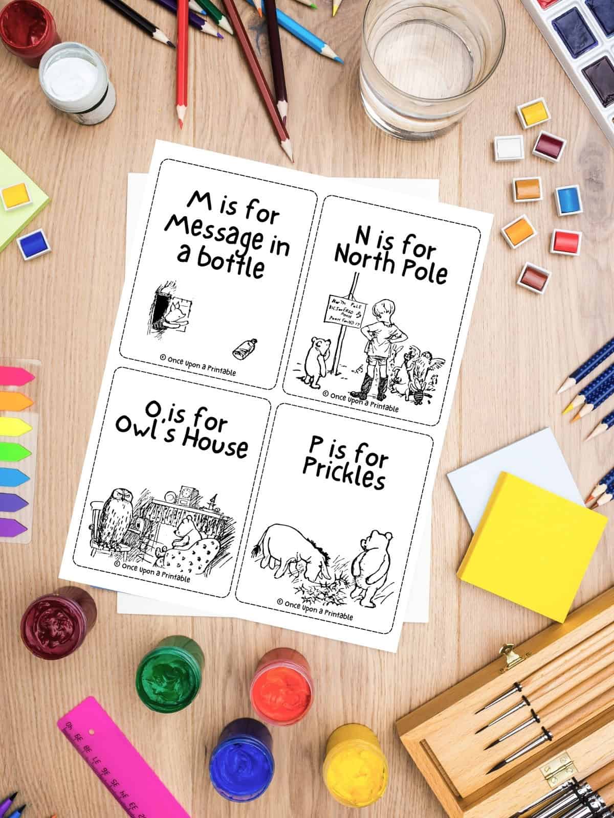 Classic Pooh Alphabet flashcard printable laying on a table with a chaotic art supply conglomerate.