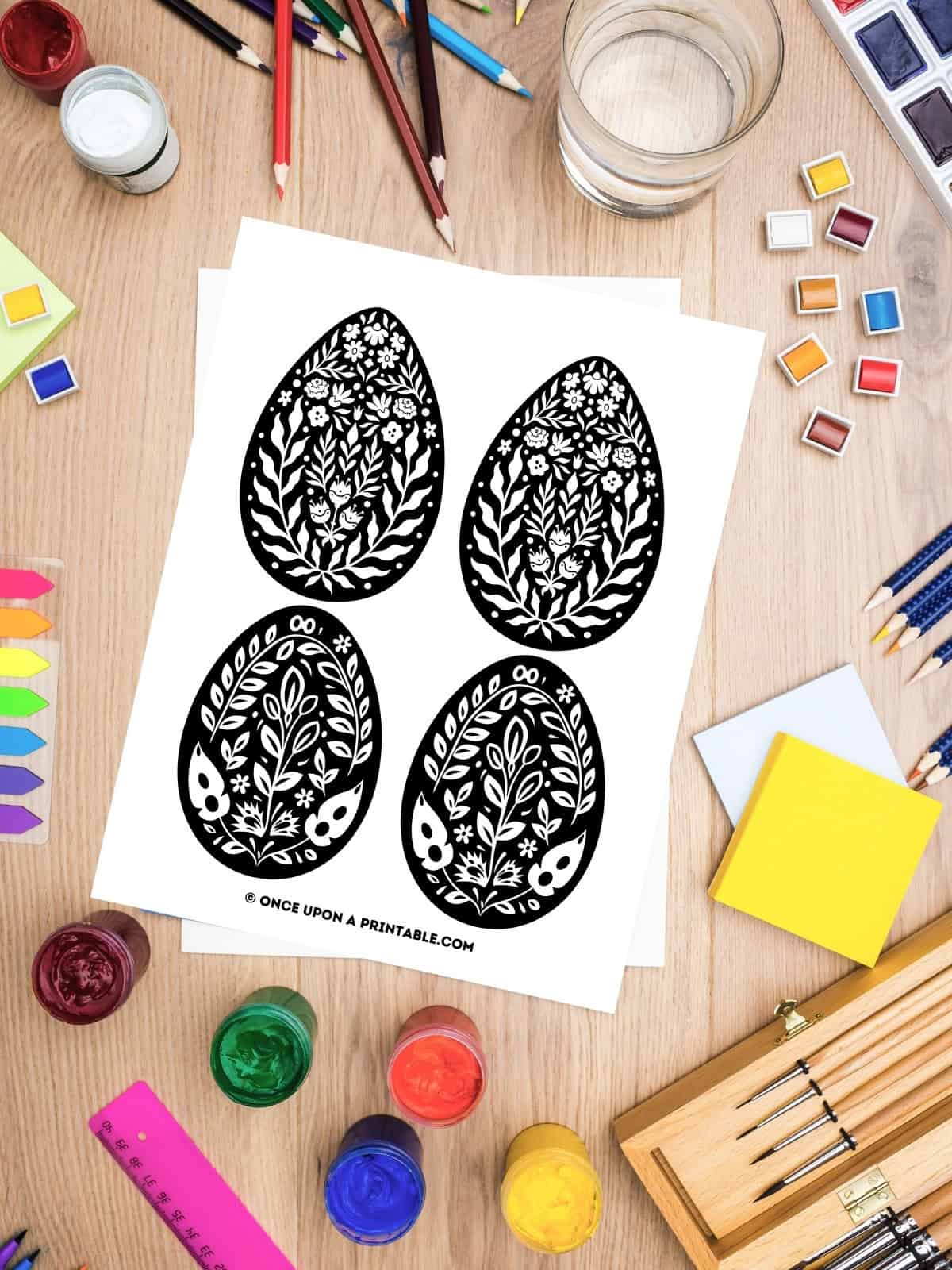 Black and white folk art Easter egg coloring pages laying on a table with coloring supplies.