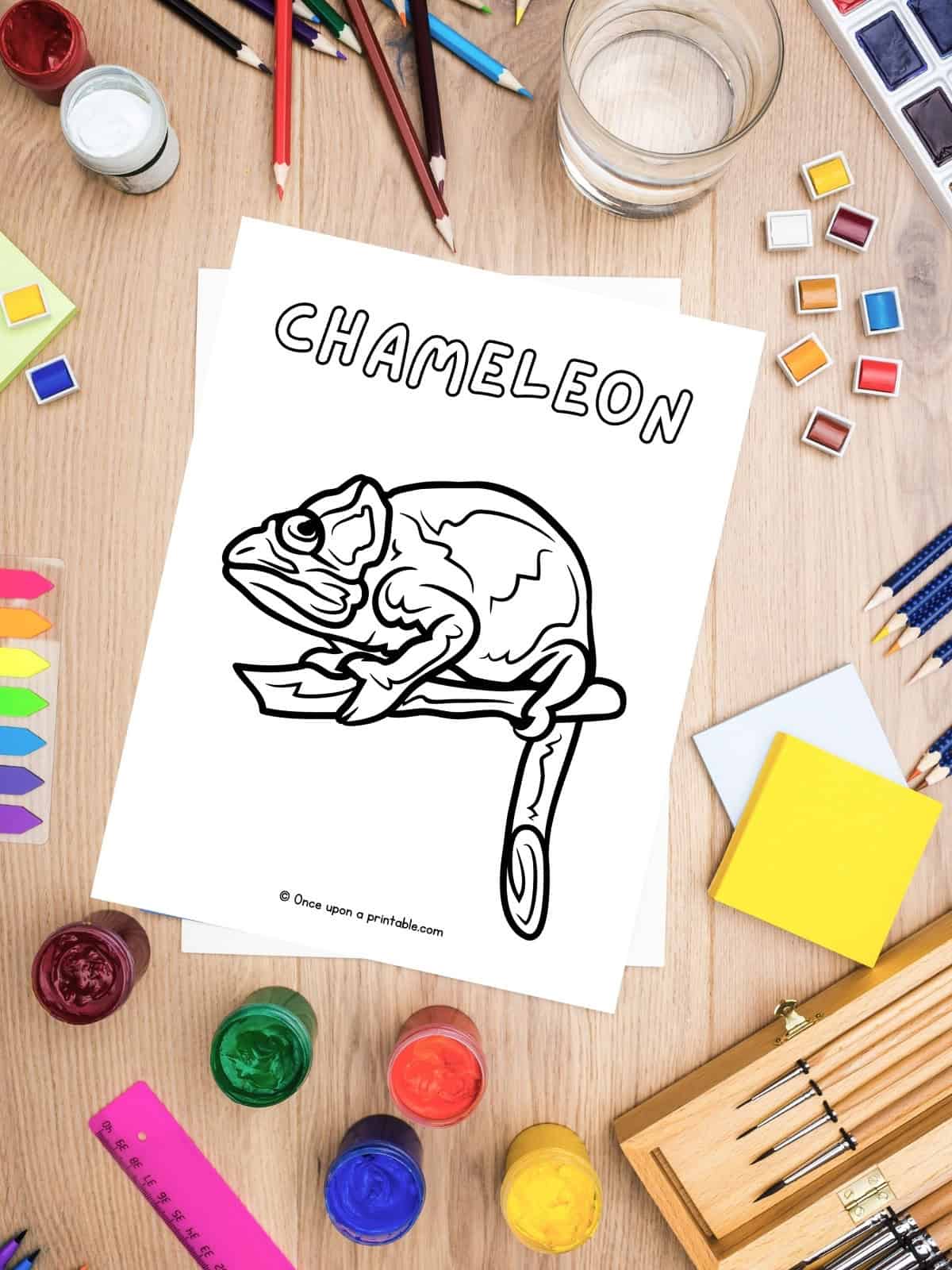 C is for Chameleon coloring page laying on a wooden table with art supplies.