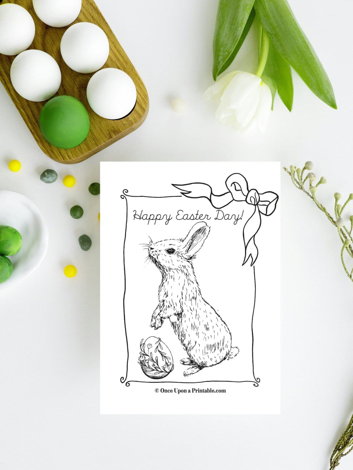 Happy Easter bunny coloring page laying on a white table with white and green eggs and a tulip.