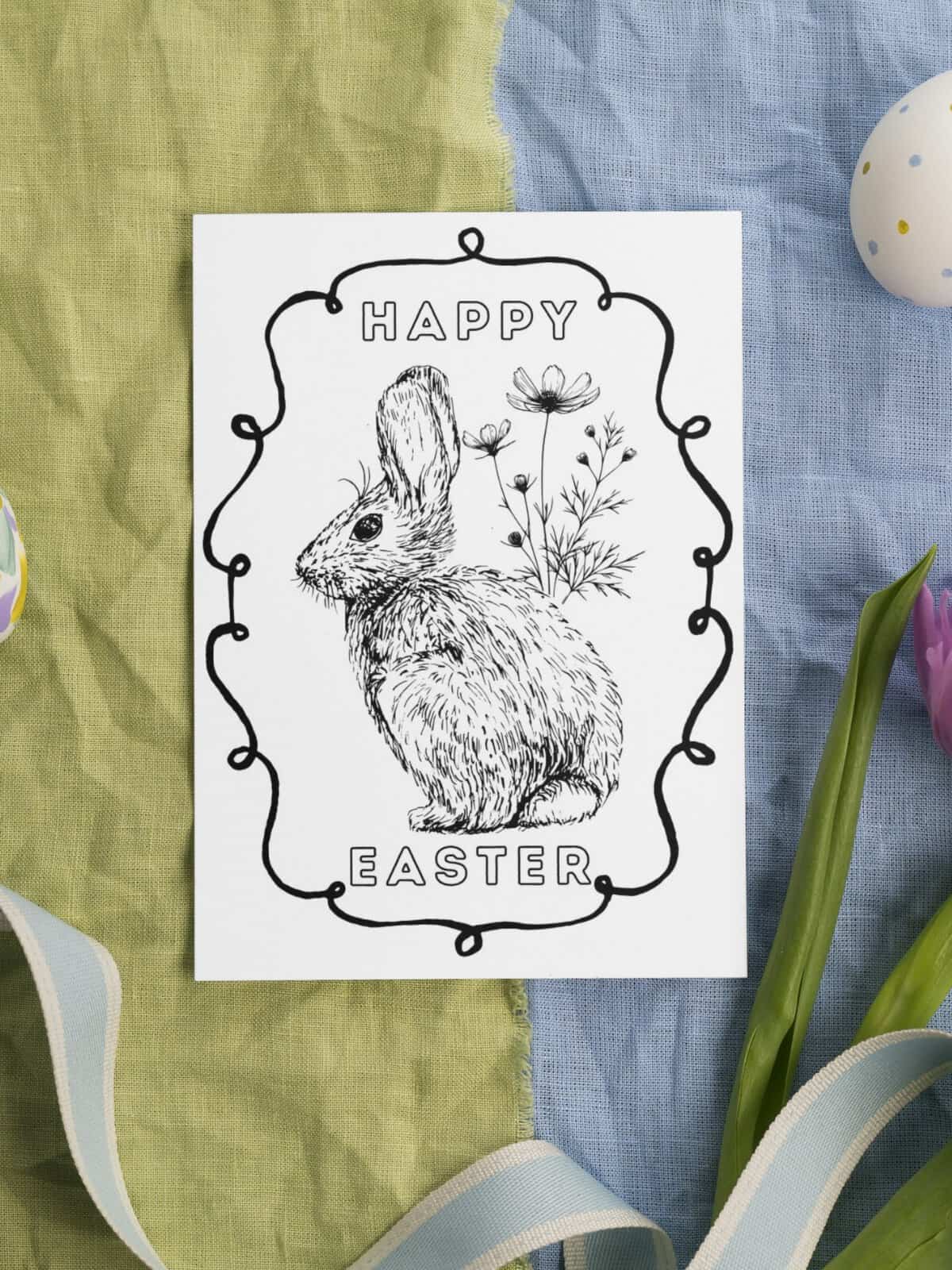 Happy Easter bunny coloring card with flowers laying on pastel fabric.