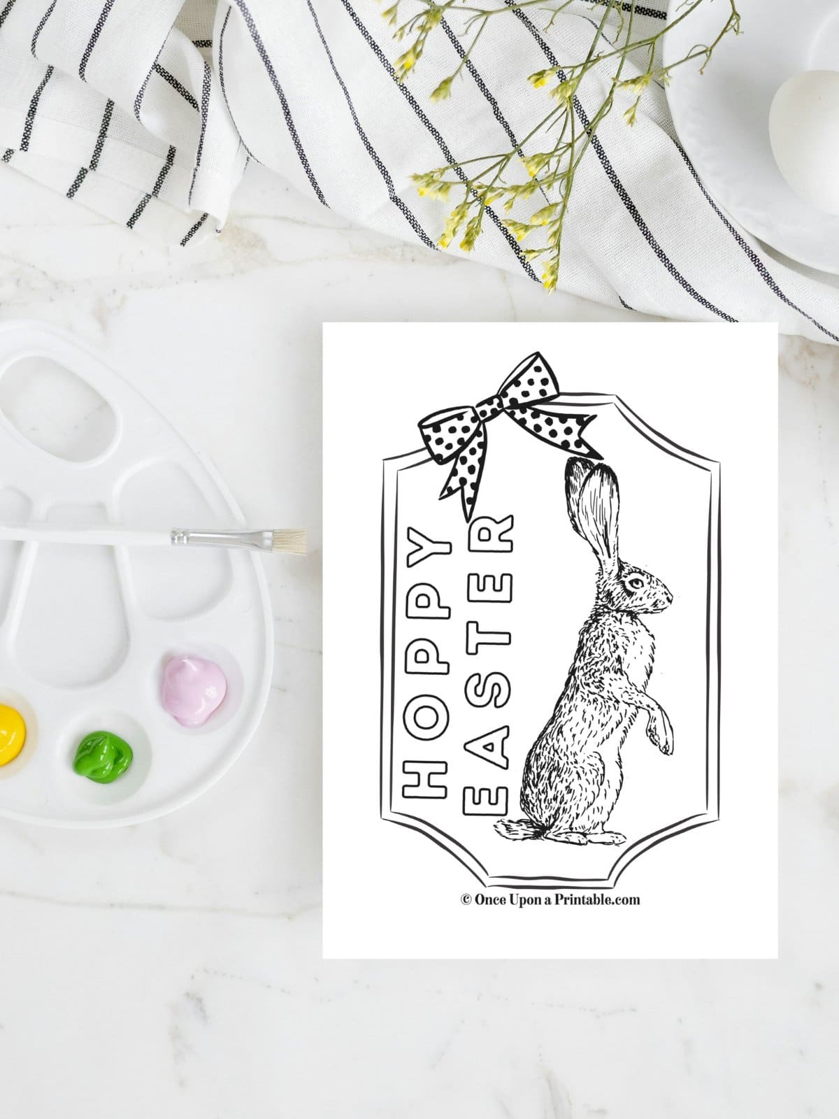 Hoppy Easter with a coloring page of the Easter hare laying on a marble tabletop with paints and a kitchen towel.