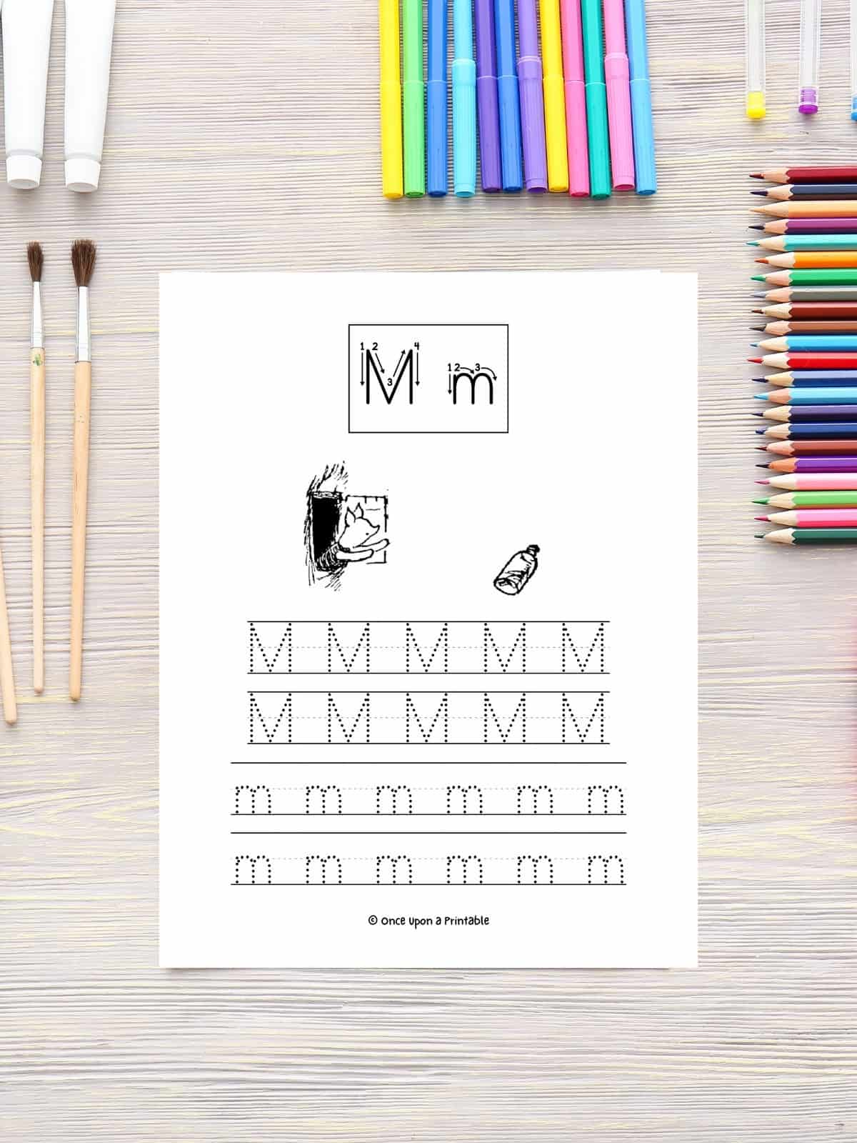 Capital M and lowercase M letter tracing worksheet with original illustrations of piglet laying on a table with coloring pencils.