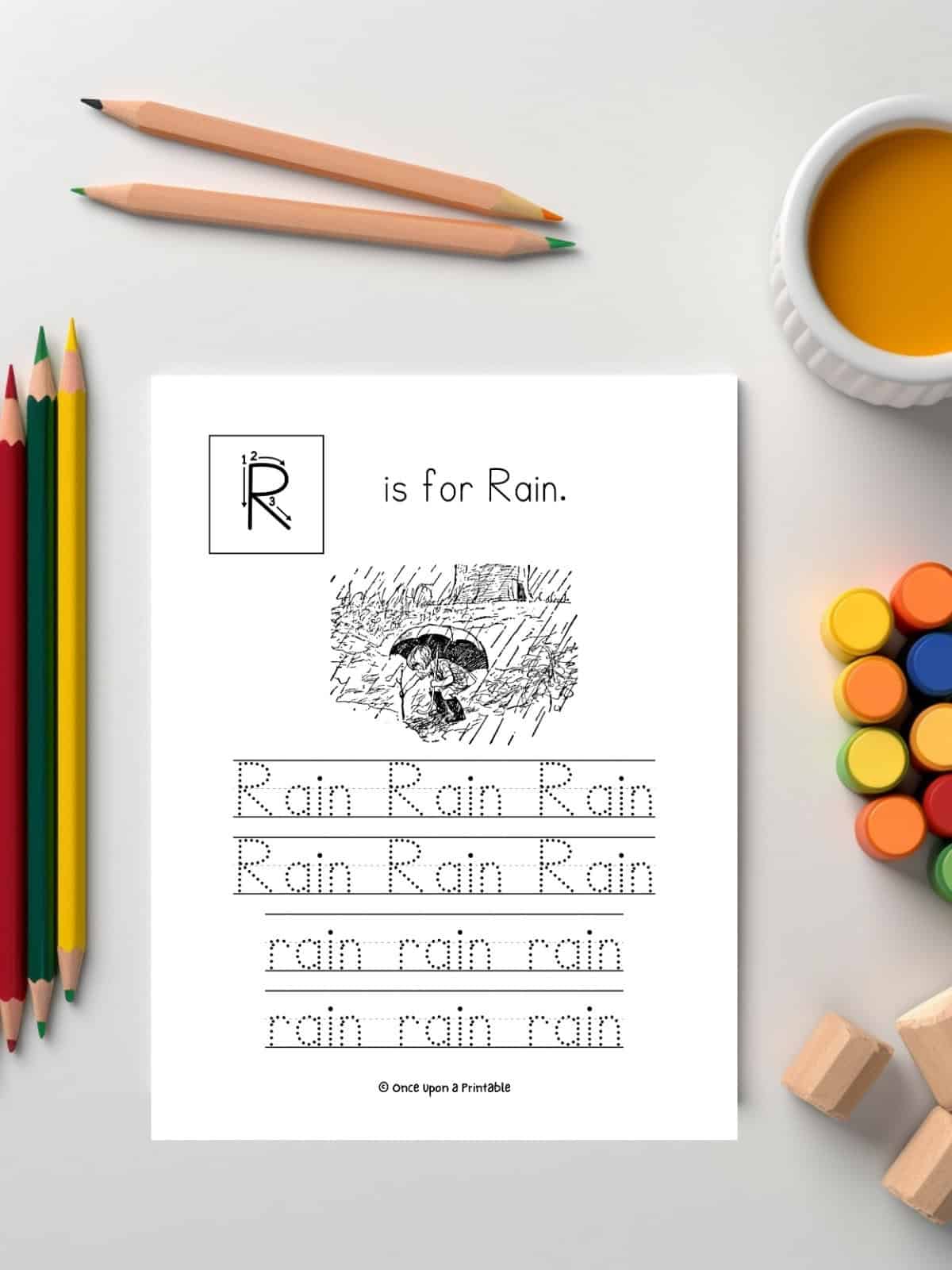 R is for rain letter and word tracing worksheet with illustrations by e. h. shepard laying on a table with coloring supplies.