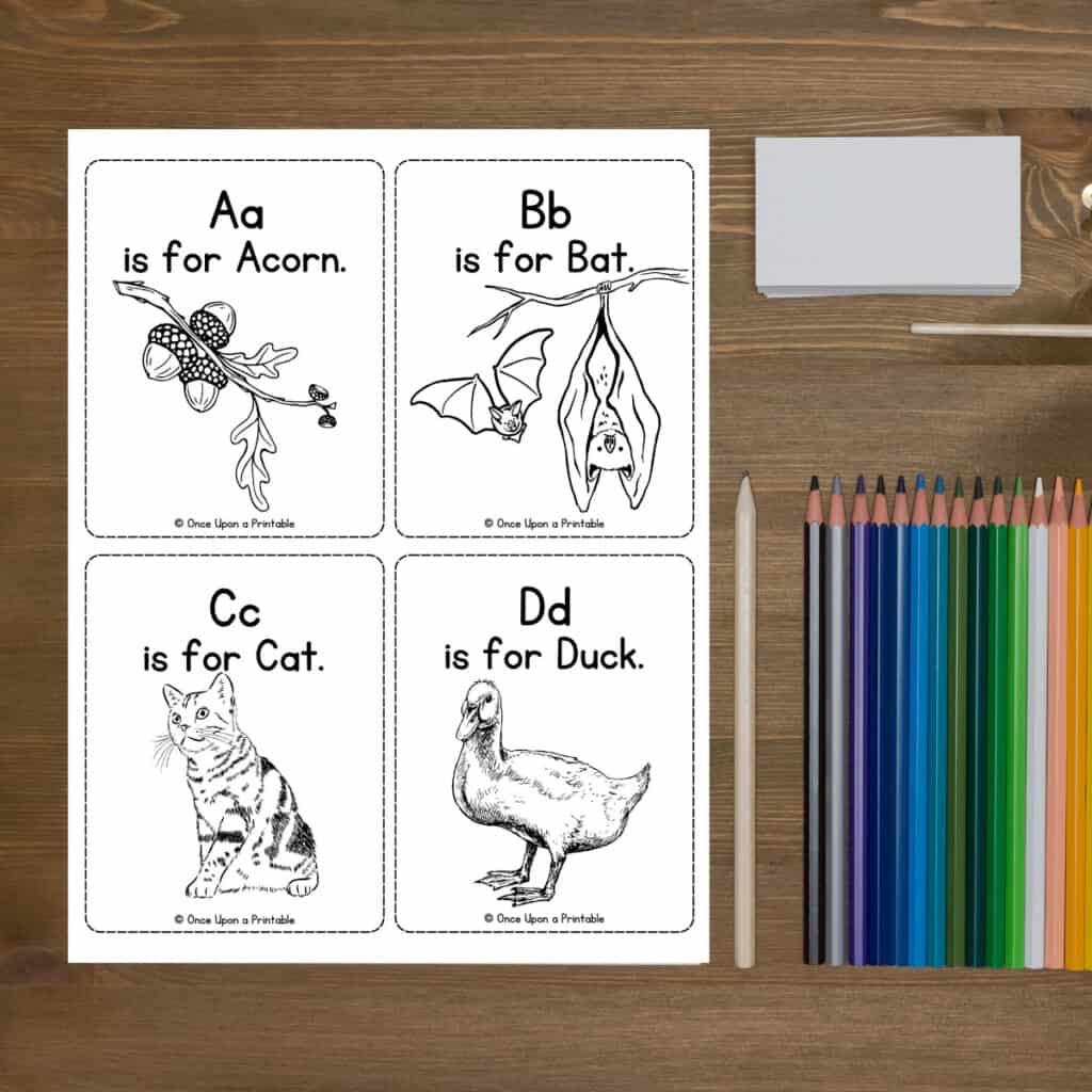 Simple alphabet cards that are in black and white coloring page laying on a wooden table with coloring pencils.